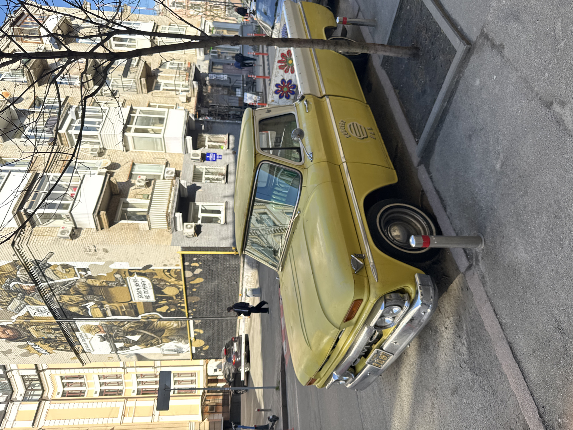 The image captures a street scene in Kyiv, Ukraine. A vintage yellow pickup truck with a white roof is parked along the sidewalk. The truck has a design on its side and flowers on the truck bed. Behind the truck, a building with multiple apartments and balconies can be seen. A mural depicting soldiers is painted on the side of the building. A few people walk on the sidewalk and there are cars on the street. A bare tree branch extends into the upper left of the frame.