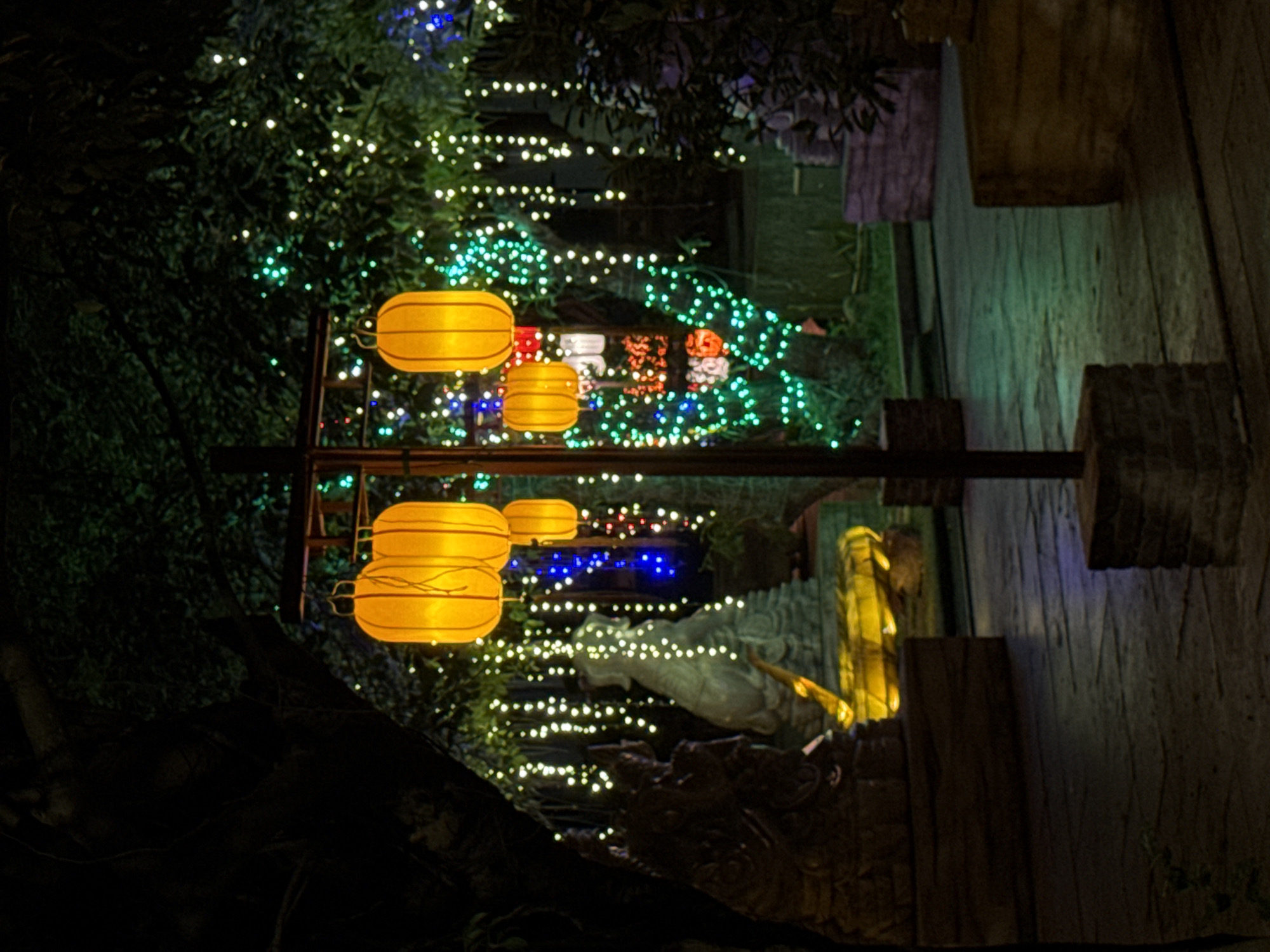 This is a nighttime photograph of a garden area decorated with numerous lights, likely in Đà Nẵng, Vietnam, given the context provided. The scene is dominated by the warm glow of several yellow, cylindrical lanterns hanging from a central pole. These lanterns are illuminated from within, casting a soft light.
Surrounding the lanterns, and scattered throughout the background, are many small, colorful lights. These appear to be strung along branches of trees and possibly bushes, creating a festive and magical atmosphere. The lights are predominantly green and white, with some blue and red lights also visible.
In the mid-ground, to the left, are two stone sculptures. The one in the foreground is dark and appears to be a mythical creature or deity, possibly with intricate carvings. Behind it is a lighter-colored, more abstract sculpture, perhaps representing a seated figure or a water feature. These sculptures are also illuminated, with some parts bathed in a yellow light.
The ground in the foreground is paved with dark, possibly wet, stone slabs. The overall environment is that of an outdoor garden or courtyard, with trees and foliage forming a dark backdrop against the illuminated decorations. The lighting creates a contrast between the brightly lit areas and the dark shadows, adding depth and drama to the scene. There are no people visible in the photograph.