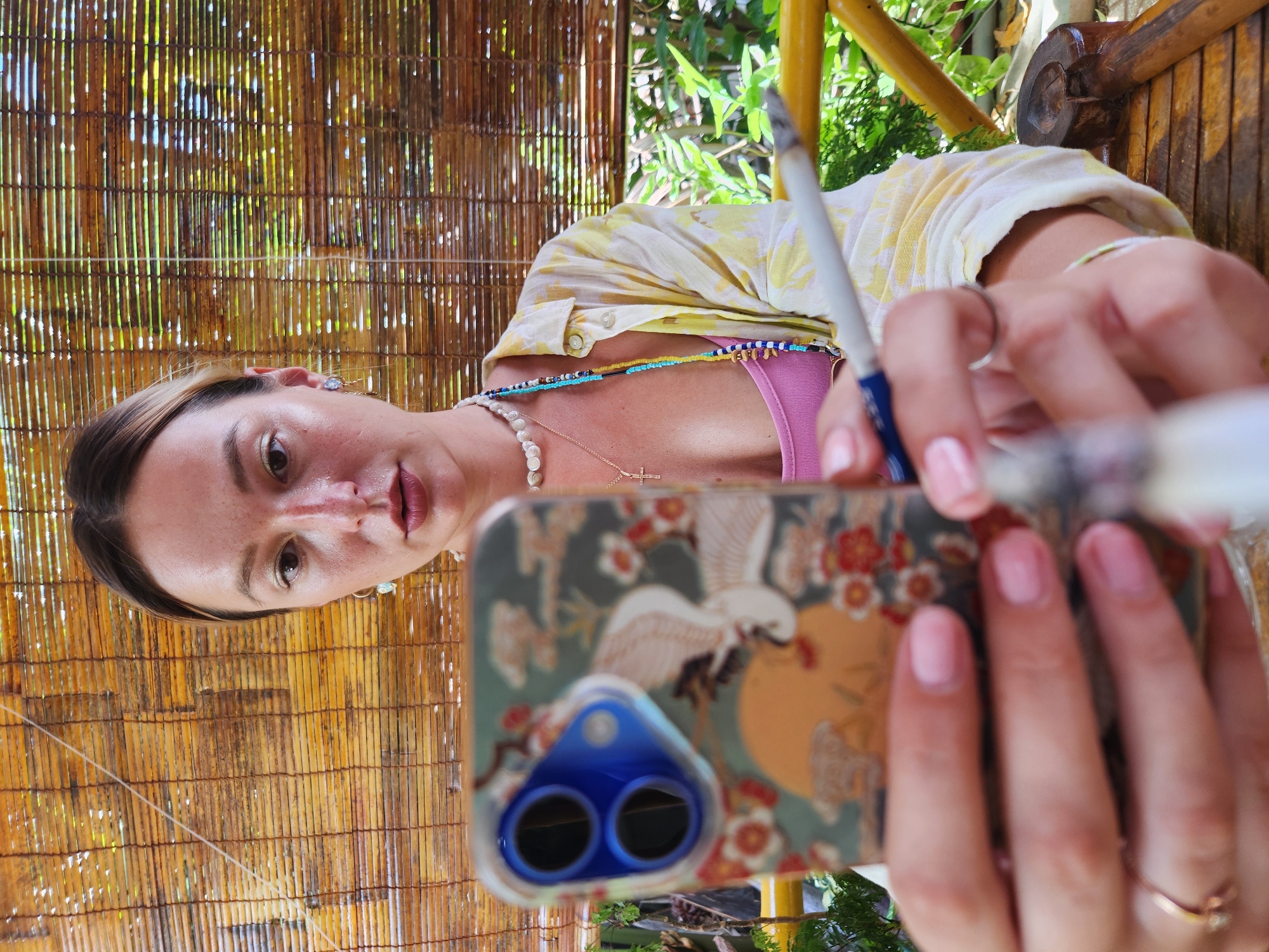 A young woman is holding a smartphone with a floral and crane design case in front of her face. She is looking into the phone's screen, and her hands are positioned to operate it. She is wearing a pink top and an open yellow and white floral patterned shirt. Around her neck, she has a pearl necklace and a beaded necklace. A cross pendant is visible on the pearl necklace. Her fingernails are painted pink. In her right hand, she is holding a slender white object with a blue tip, possibly a pen or a makeup brush. The background consists of a bamboo blind, suggesting an outdoor or semi-outdoor setting, possibly a cafe or a balcony. Lush green foliage is visible behind the bamboo blind, and a yellow railing is present to the right. A wooden object, possibly a tool, is partially visible on a wooden surface in the lower right corner. The image has a shallow depth of field, with the woman's face and hands in sharp focus and the background blurred. The lighting suggests daylight. The scene appears to be set in Da Nang, Vietnam, given the context provided.
