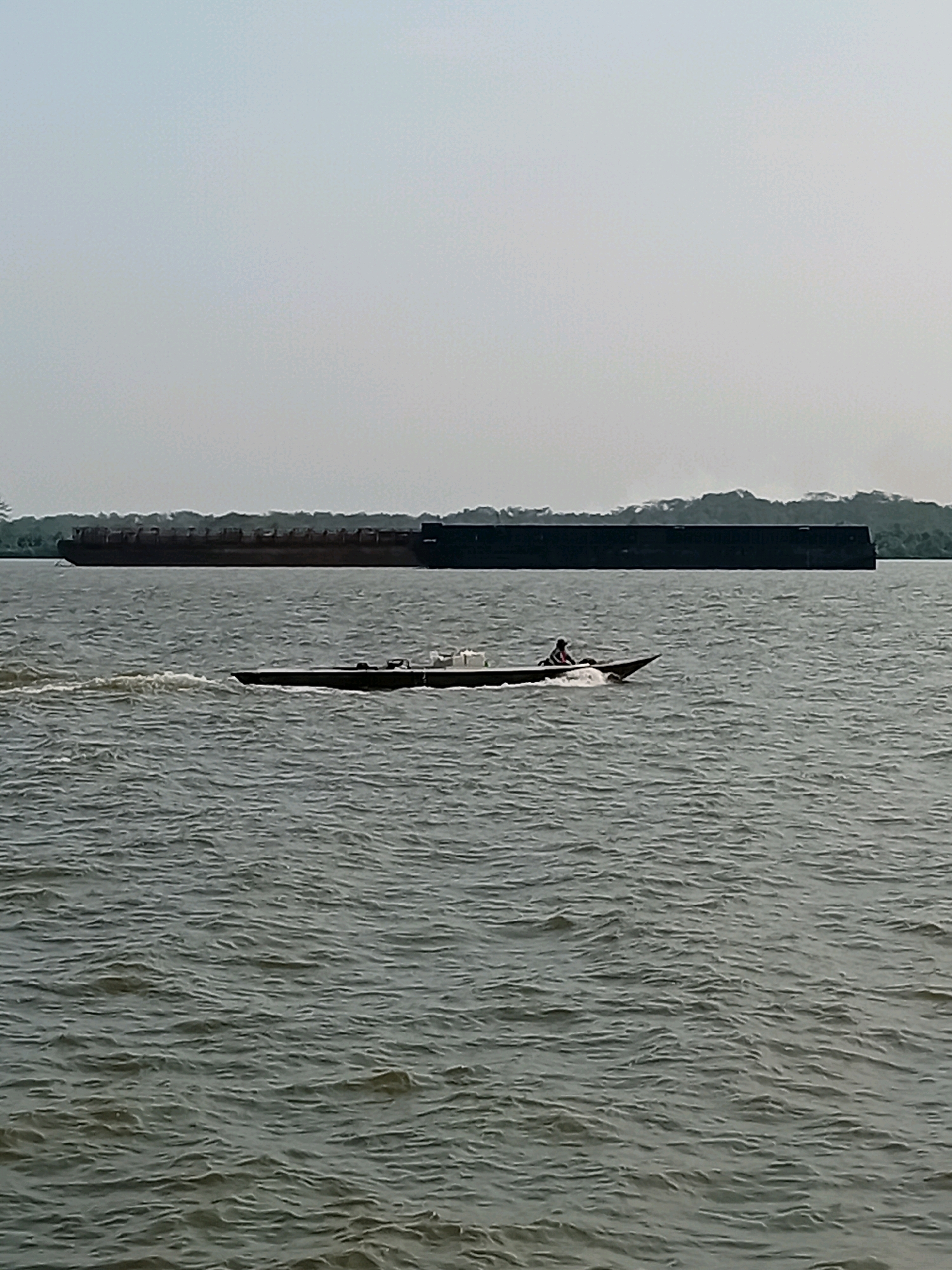 In Sungai Meriam, Indonesia, a long, dark barge is docked in the distance on a wide body of water. In the foreground, a small, dark boat with a person at the helm speeds across the choppy, grey water, leaving a wake behind it. The sky is overcast and hazy, with a faint outline of trees visible along the distant shoreline.