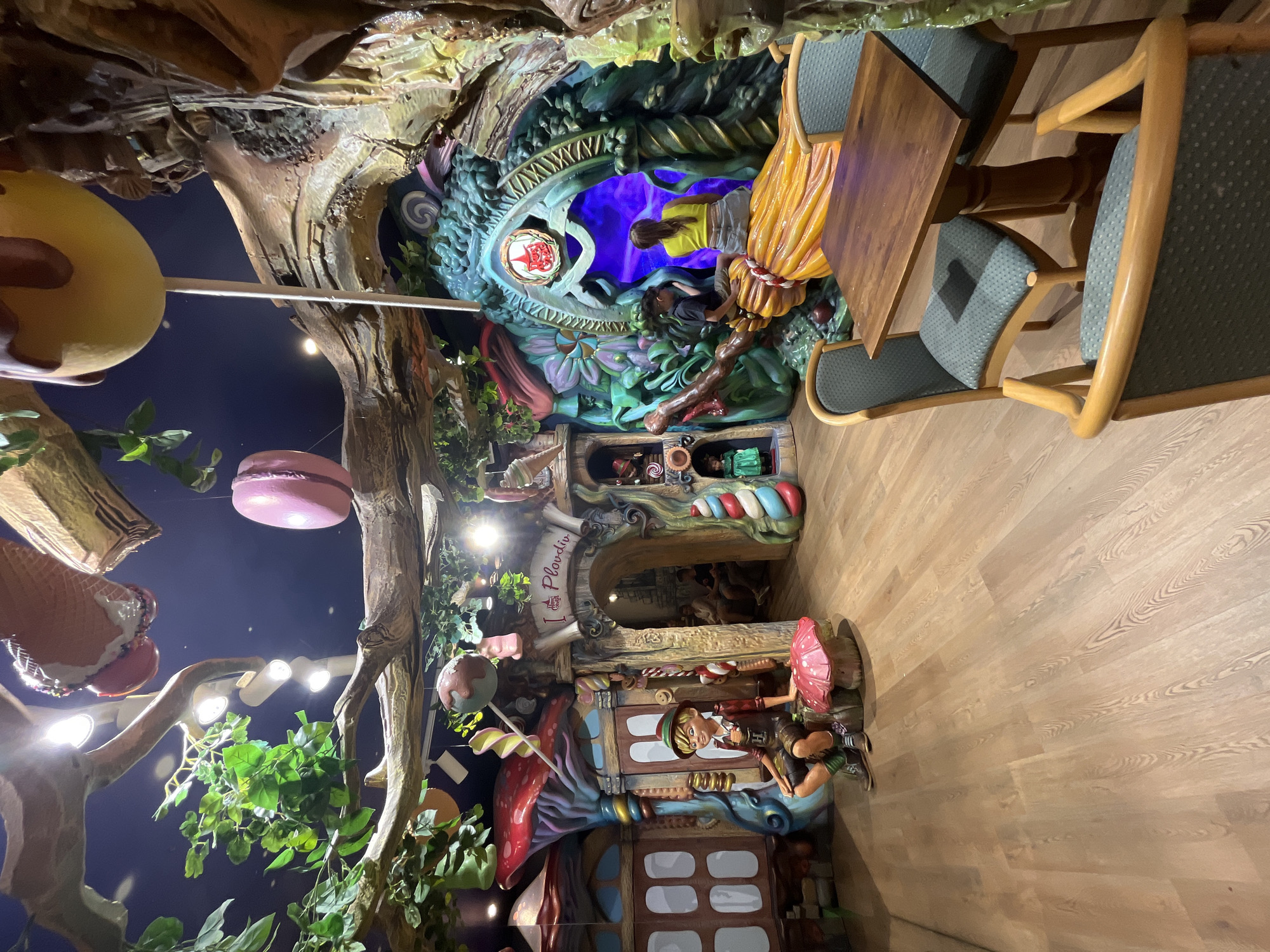 This image depicts the whimsical interior of a themed establishment, identified by a sign displaying "I Love Plovdiv" above an archway, placing the location in Plovdiv, Bulgaria. The environment simulates an enchanted forest or fairytale setting, characterized by oversized, vibrant decorations.
The ceiling features faux tree branches intertwined with foliage and integrated overhead lights. Several large, confectionery-themed objects, including a pink macaron, lollipops, and what appears to be a melting ice cream cone or candy, are suspended from the branches.
On the left, a structure resembling a house with a large red mushroom cap roof is visible. In front of it, a statue of a young boy, reminiscent of Pinocchio, dressed in traditional attire with a feathered cap, sits on a smaller red mushroom. To the right of this statue, a column decorated with red and white candy cane stripes ascends.
In the center, an archway with a sign "I Love Plovdiv" leads into a dimly lit interior where several individuals are partially visible.
To the right, a large, intricate structure designed to resemble gnarled roots, vines, and fantastical foliage forms another archway. Inside this archway, a luminous purple light emanates. A substantial, golden-yellow sculpture resembling a giant broomstick extends from the lower part of this structure. An adult female wearing a yellow top and a child are seated on this broomstick, facing into the glowing purple area. Adjacent to this structure are small recessed niches containing dolls or figures.
The floor is composed of light-colored wooden planks. In the foreground, two small, square wooden tables are present, each accompanied by two chairs upholstered in a light blue fabric with a subtle pattern. One table and its chairs are prominently visible on the right, unoccupied.