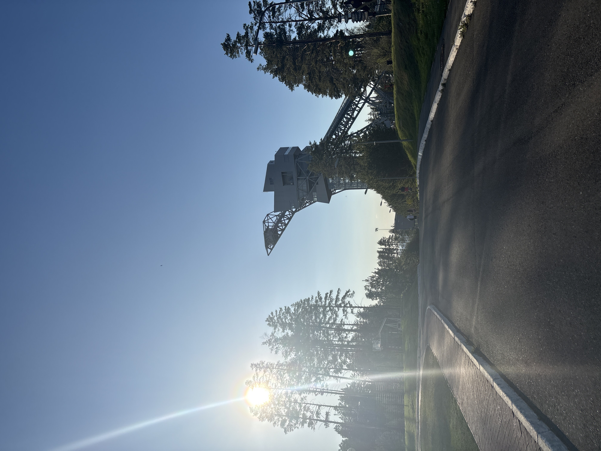 The image shows a view in Прикамский, Russia. A large, gray metal crane structure is seen in the upper-center, partially obscured by trees. Below the crane, the sun is shining brightly behind a row of trees, creating a lens flare effect. The foreground shows a paved walkway with a concrete curb, adjacent to a dark asphalt road. The sky is clear and blue.