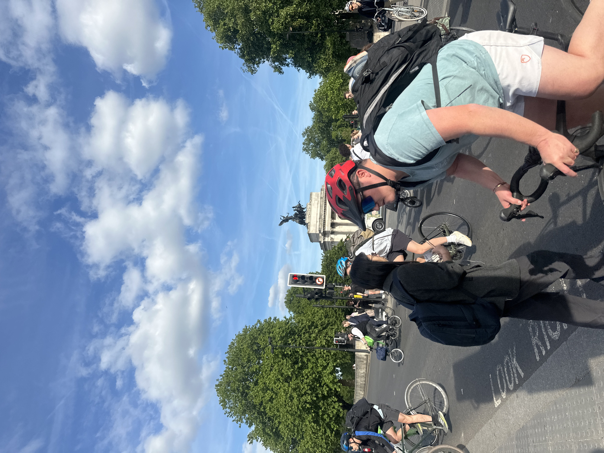 The image depicts a street scene in London, United Kingdom. Several cyclists are stopped at a red traffic light near a pedestrian crossing marked "LOOK RIGHT". The sky is blue with scattered clouds. In the background, the Victoria Memorial is visible, suggesting the location is near Buckingham Palace. People are either on bicycles or standing, waiting for the light to change. The image is taken from a slightly elevated perspective.