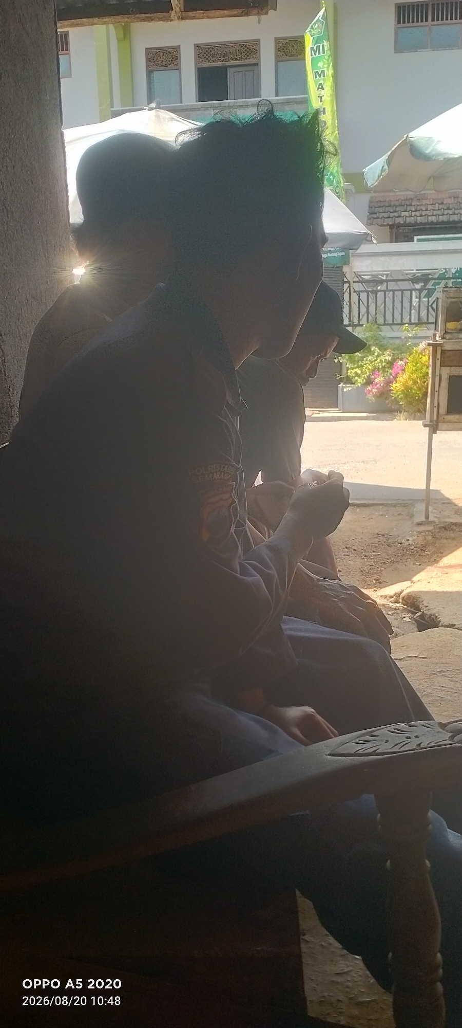 The image shows two individuals sitting on a wooden chair outside a building in Bugel, Indonesia. The person in the foreground, wearing a dark uniform with a patch that appears to say "POLRI" and "SEMARANG," is partially obscured by shadow and has their head turned to the right, looking down. The second person, seated to their left and also facing right, is less visible. In the background, a white building with green trim and intricate window carvings is visible, along with a green flag bearing text. The environment appears to be a street or outdoor seating area, with sunlight casting shadows and illuminating the scene. There are also decorative elements on the building and the surrounding area, including potted plants. The date stamp on the image indicates it was taken on August 20, 2026, at 10:48 AM.
