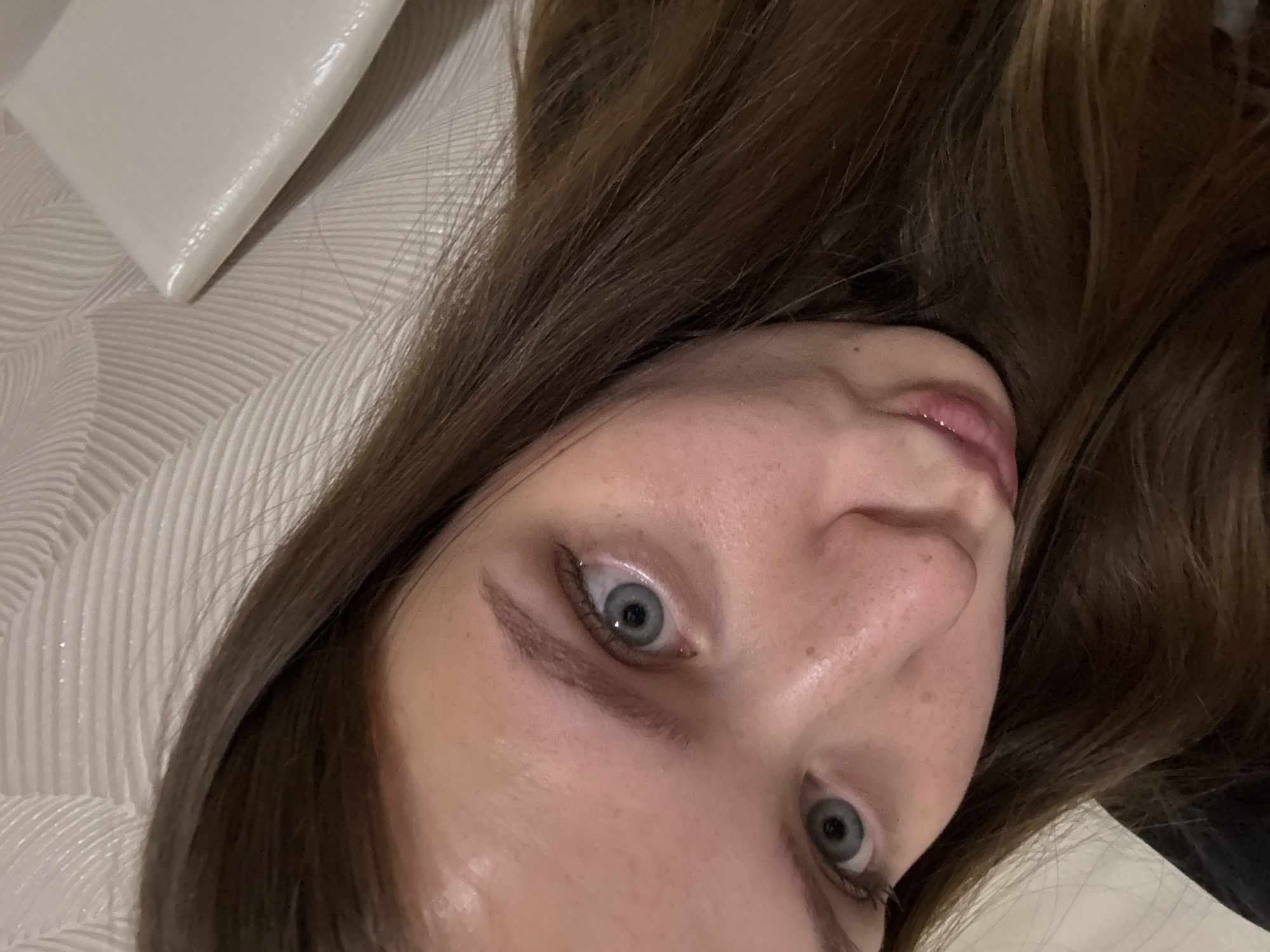 The image displays a close-up, slightly tilted portrait of a young person, likely female, situated indoors within the context of Chaikovsky, Russia. The subject has fair skin, with visible freckles scattered across her nose and cheeks, and striking light blue-grey eyes that gaze directly towards the viewer. Her long, wavy brown hair frames her face and spreads across a white, textured surface, which appears to be a pillow or bedding with a subtle pattern of curved, raised lines.
The person seems to be lying down, potentially taking a selfie or being photographed from an overhead angle. Her expression is neutral, possibly curious or relaxed, with her lips slightly parted. Subtle eye makeup, including mascara and eyeliner, enhances her eyes. The lighting is soft and consistent with an indoor setting. No specific time of day or weather conditions are discernible, and there is no visible text in the media file. The overall impression is of an intimate, casual moment captured indoors.
