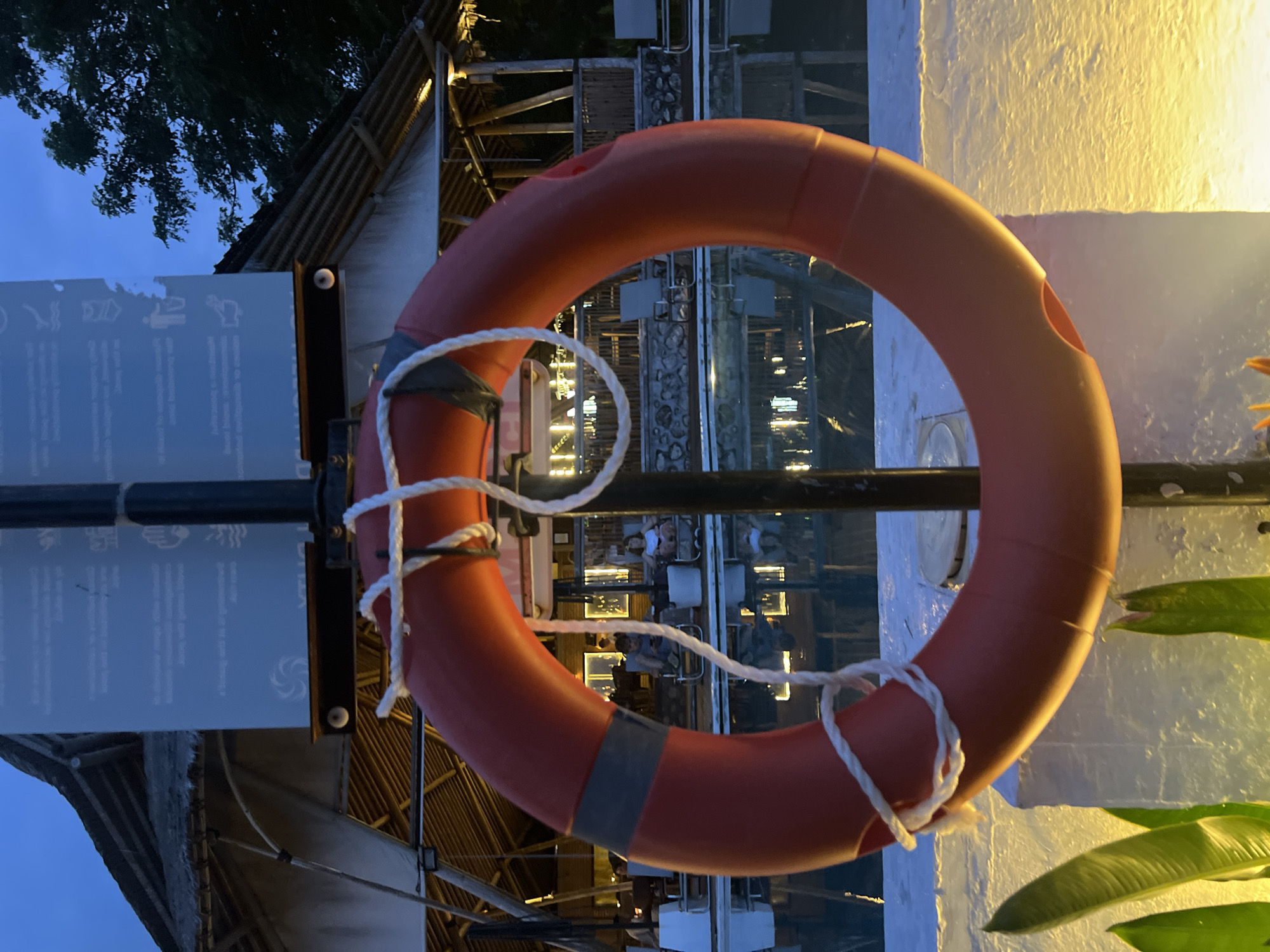 A prominent red lifebuoy with white rope accents hangs in the foreground of this image, appearing to frame the scene beyond. It is suspended from a dark metal pole, which also supports a translucent sign to its left. This sign features several faint white icons, including symbols for a swimmer, waves, a hand, a person with a dog, and a circular swirl, along with indistinct text, likely indicating safety regulations or instructions.
Through the opening of the lifebuoy and behind the sign, a multi-level building is visible, possibly a restaurant or resort. Its structure incorporates wooden beams and some stone-like textures. Inside, soft artificial lighting illuminates the space, where blurred figures of people can be seen seated at tables, suggesting an evening dining or social setting.
The upper left corner reveals a dark blue sky with silhouettes of tree branches, indicating the time of day is dusk or night. To the right of the lifebuoy, a white wall is brightly lit by a warm yellow light emanating from an unseen source, adding to the evening ambiance. Green foliage is partially visible in the bottom right, contributing to a lush, tropical feel. The scene conveys a calm and inviting atmosphere, typical of a waterside establishment in a location like Nusa Penida, Indonesia, during the evening hours.
