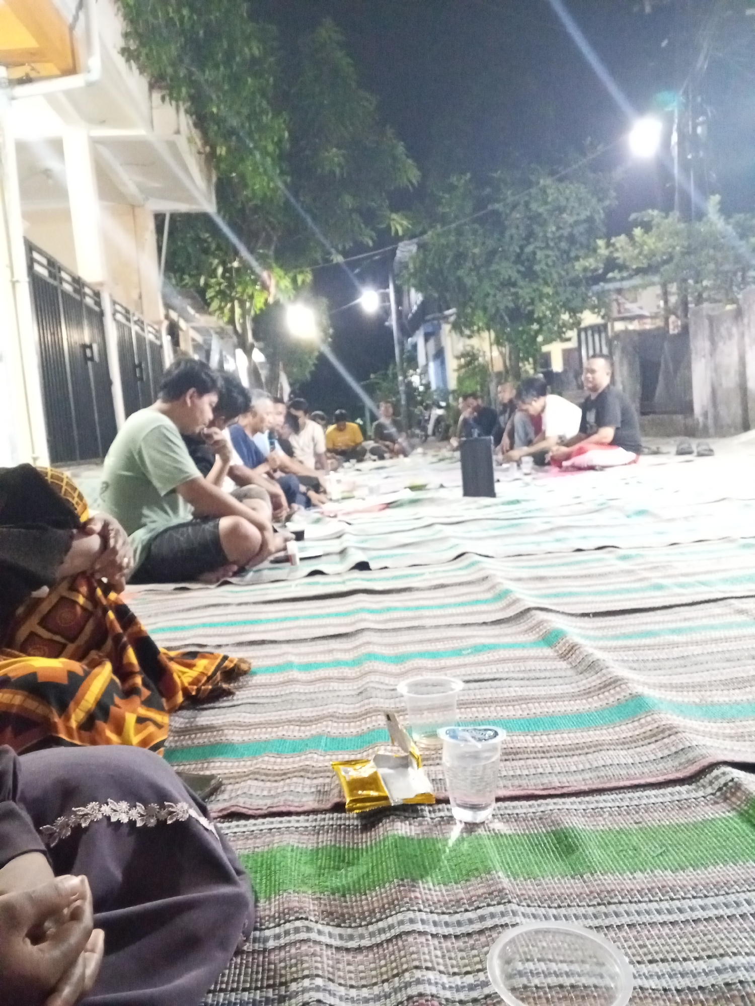 In Ciruas, Indonesia, a group of men are gathered outdoors at night, sitting on striped mats spread on the ground. The scene appears to be a casual social gathering, possibly a meal or a religious observance, given the time of night and the communal seating arrangement. The men are dressed in casual clothing, including t-shirts and shorts. Some are looking at each other, suggesting conversation or interaction. In the foreground, there are plastic cups and what appears to be a snack packet. The background shows the facade of a building with lights and trees, indicating an urban or semi-urban environment. The overall atmosphere is relaxed and informal.