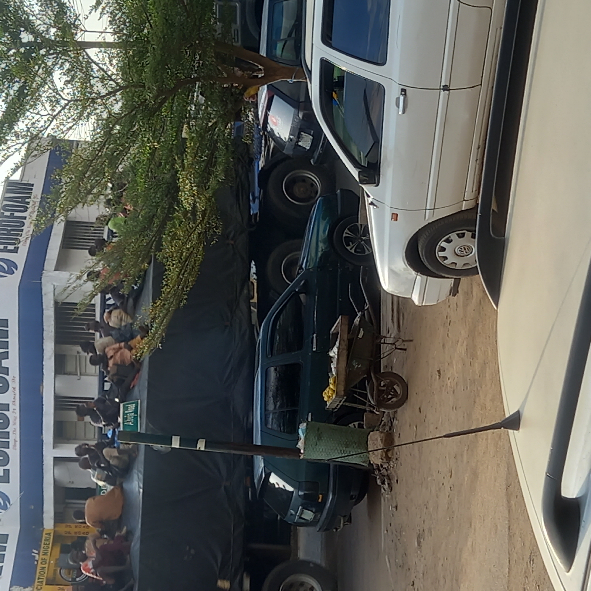 The image depicts a scene in Sardauna, Nigeria, adjacent to a EUROFOAM store. A large group of people are seated on a platform covered in black tarpaulin, near a street sign that reads "Abuja Road". Above the platform and crowd, the EUROFOAM building is visible with its blue and white signage. Several vehicles are parked on the road, including what appears to be a truck loaded with additional vehicles, a green sedan, and a white van. A wheelbarrow is near the vehicles, and a tree's branches extend over the scene. The image is shot at ground level and appears to be rotated or shot from an angle.