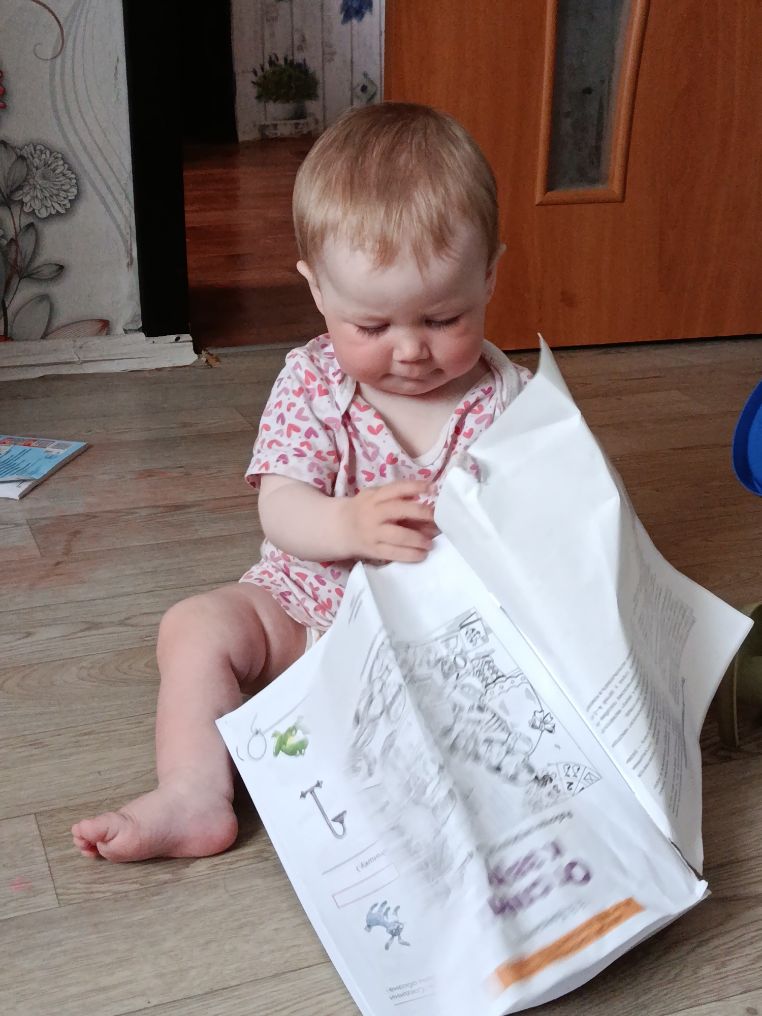 A child sits on a wood-look floor in a room with floral wallpaper and a wooden door, opening a large white paper with illustrations and text. The paper has a drawing of a cat, a "T" with the word "Оборотень" underneath, and a drawing of a blue creature at the bottom. The child is wearing a white and pink heart-patterned shirt. The city of Фоки, Russia, is not discernible from this image.