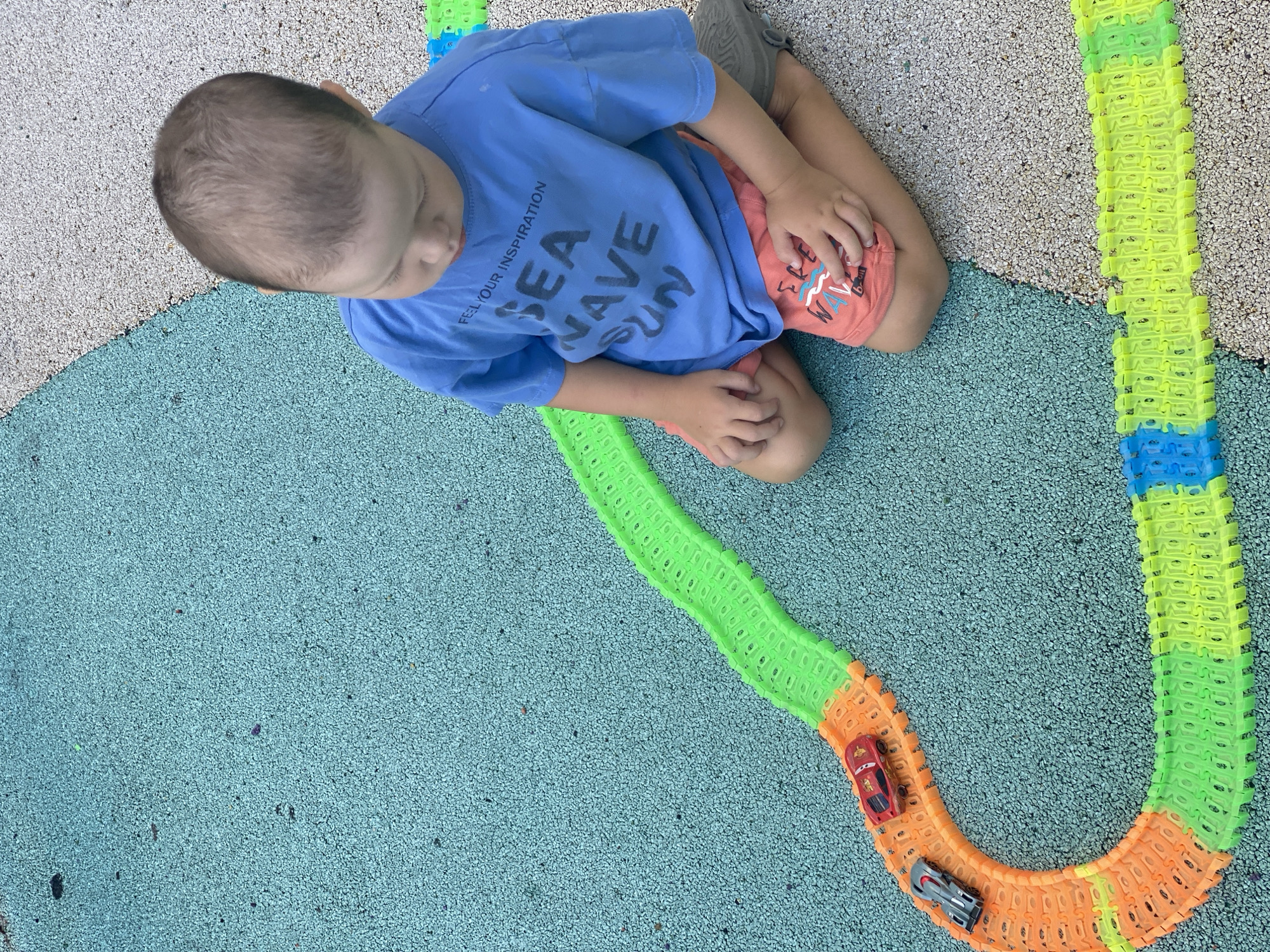 A young child, possibly a toddler, kneels on a textured, light-blue surface, facing away from the camera and looking down at a toy race track. The track, made of interlocking plastic segments in orange, green, and blue, winds across the surface. Two toy cars, one red and one gray, are positioned on the track. The child wears a blue t-shirt with the words "FEEL YOUR INSPIRATION SEA WAVE SUN" printed on it in dark lettering, and orange shorts. The surface transitions to a light gray, gravel-like material. The background is blurred, suggesting an outdoor setting. The image does not provide enough context to determine a specific location in Sochi, Russia.