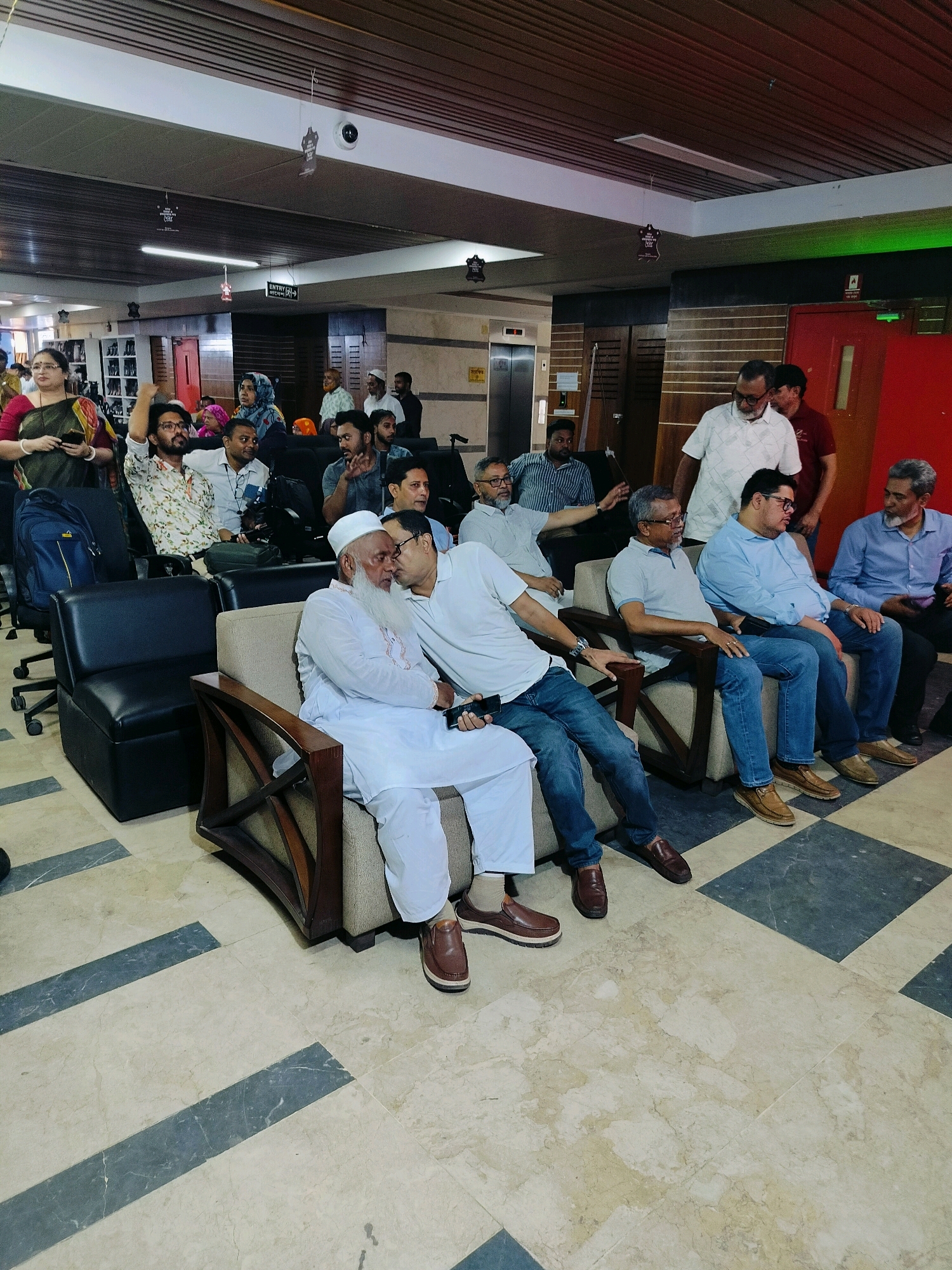 In a well-lit indoor space, likely a reception or waiting area, a group of people are seated and standing. The architecture suggests a modern building, possibly in Dhaka, Bangladesh, with a dropped ceiling and tiled flooring in a beige and grey checkerboard pattern.
In the foreground, two men are seated on a sofa. The man on the left, wearing a white thobe and a white cap, has a long white beard and appears to be engaged in conversation. The man to his right, wearing a light blue shirt and dark jeans, is leaning in and speaking to him, holding a mobile phone in his left hand.
To the left of the sofa, a man in a white patterned shirt and dark jeans is seated in a black chair. He is looking upwards and to his left, with his right arm raised. Next to him, a person in a green and red sari is standing, holding a mobile phone in their left hand and looking to the left.
Further back, several other individuals are visible. A group of men in white shirts and various colored trousers are seated on chairs and sofas. Some are looking forward, while others are interacting amongst themselves. One man in a white collared shirt is standing and looking towards the group seated on the sofas. In the background, an elevator door is visible. Overhead, there are ceiling lights and decorative elements, including what appear to be small festive ornaments. The overall atmosphere seems to be one of a casual gathering or a break during an event.