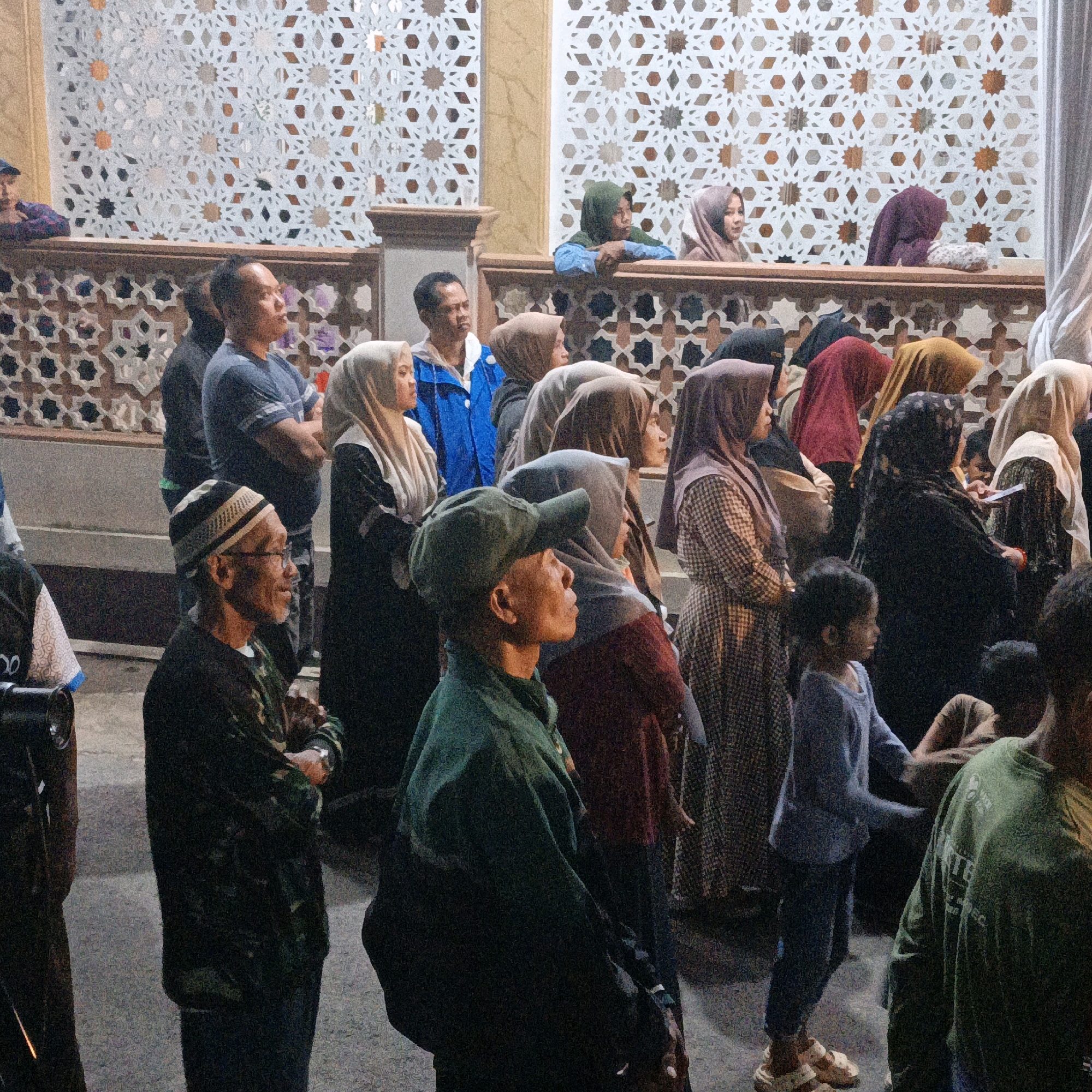 The image captures a nighttime outdoor gathering in the city of Kalitorong, Indonesia, likely near a mosque given the architectural style. A crowd of people, predominantly women wearing hijabs and men in casual attire, are visible. The background features intricate decorative panels with geometric patterns, typical of Islamic architecture. In the foreground, several individuals are looking in different directions, some engaged in conversation or observing the surroundings. One man in the foreground wears glasses and a cap, while another, to his right, is in a green military-style jacket. A child in a blue shirt and sandals stands towards the right side of the frame. The overall lighting suggests an ambient, possibly artificial, illumination.