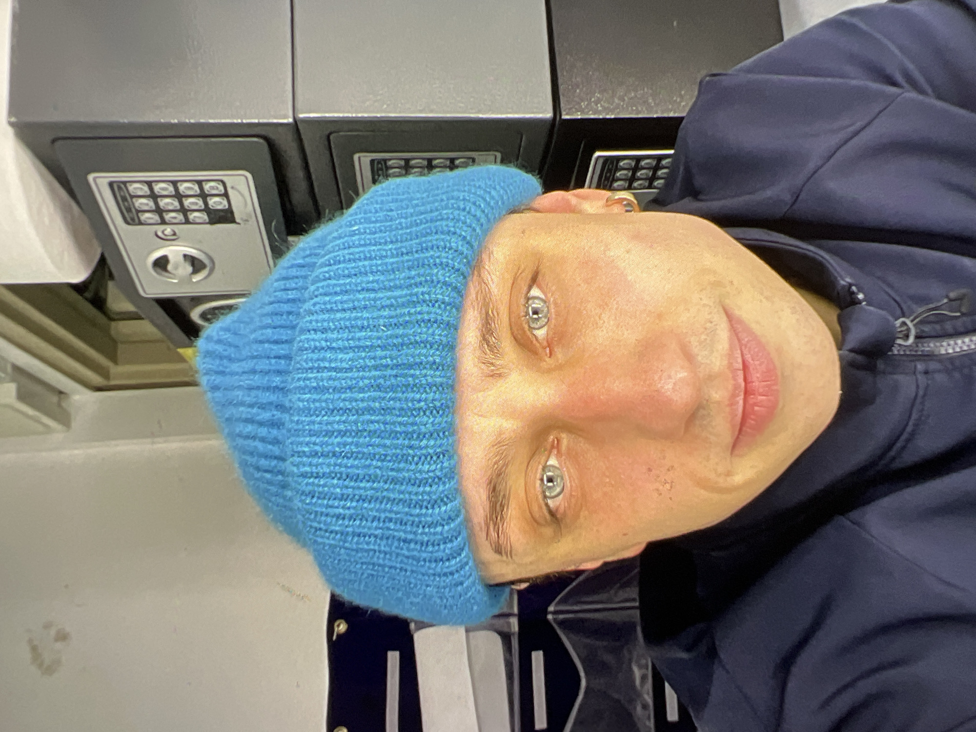 A close-up selfie of a person wearing a bright blue knitted beanie and a dark blue jacket. The person has light skin, light blue eyes, and appears to be in their late 20s or early 30s. They have a slight smile and a few freckles or moles on their cheeks. In the background, there are two stacked safes with digital keypads and a few dark blue storage organizers. The setting appears to be an indoor space, possibly a storage unit or office. There is no indication of the city of Verona, Italy.