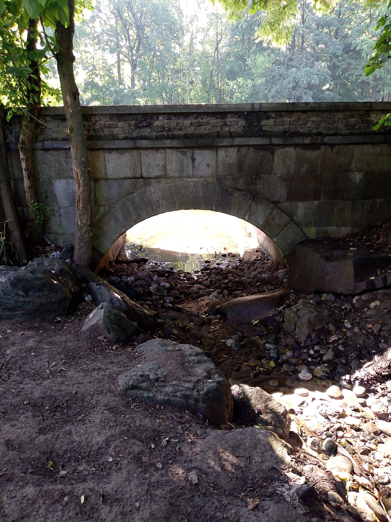 The image depicts a stone bridge arching over a shallow, rocky streambed. Trees with green foliage are visible on either side of the bridge, with a denser forest in the background. The lighting suggests it is daytime, with sunlight filtering through the trees and illuminating parts of the streambed. The bridge itself appears to be constructed from rough-hewn stone blocks. The foreground consists of a dirt path and scattered rocks. No people or animals are visible in the image. The scene appears to be in a natural park or wooded area.