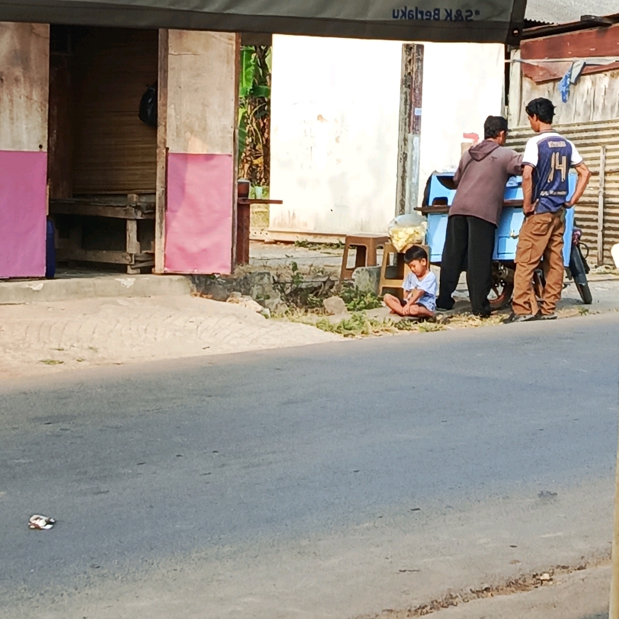 A group of three people, two adult males and one young boy, are visible near a blue street vendor cart in Karawang, Indonesia. The young boy sits on the ground, facing left and looking down. One adult male, wearing a brown hooded jacket and black pants, stands behind the cart, facing the boy. The other adult male, wearing a blue and white shirt with the number 14 on the back and brown pants, stands to the right of the cart, facing forward. The street vendor cart has wheels and appears to be selling snacks, possibly chips, which are visible in a bag on the left side of the cart. The scene is set on a paved road with a sandy area to the right, where the people and cart are located. The background consists of a building with a corrugated metal roof and wooden panels, and some greenery. A piece of litter, possibly a crumpled plastic bag or wrapper, lies on the road in the foreground. The lighting suggests it is daytime.
