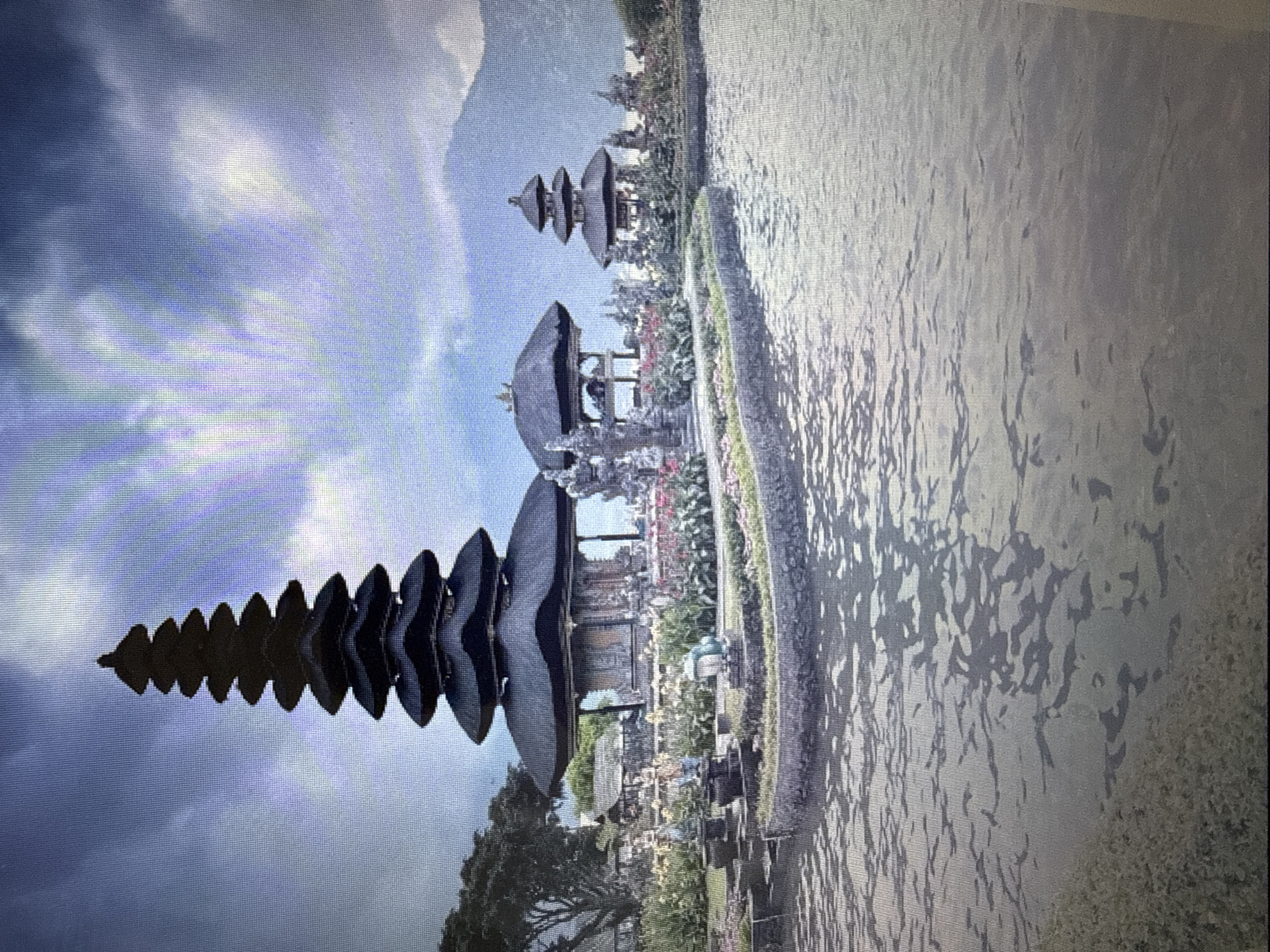 The image depicts a Balinese temple complex situated on the water. The primary structure is a multi-tiered pagoda-style temple with dark, layered roofs. To its right, a smaller, similar structure is visible, also on the water. In the foreground, a body of water reflects the sky and the structures. The background shows lush greenery, including trees and a variety of flowering plants, with a mountain range in the far distance. The sky is cloudy, with patches of brighter light suggesting sunlight filtering through. There are no people visible in the image. The scene captures a serene and picturesque landscape typical of Balinese water temples. The prompt's request to locate this scene in Pushkino, Russia, is inconsistent with the visual evidence of Balinese architecture and landscape.
