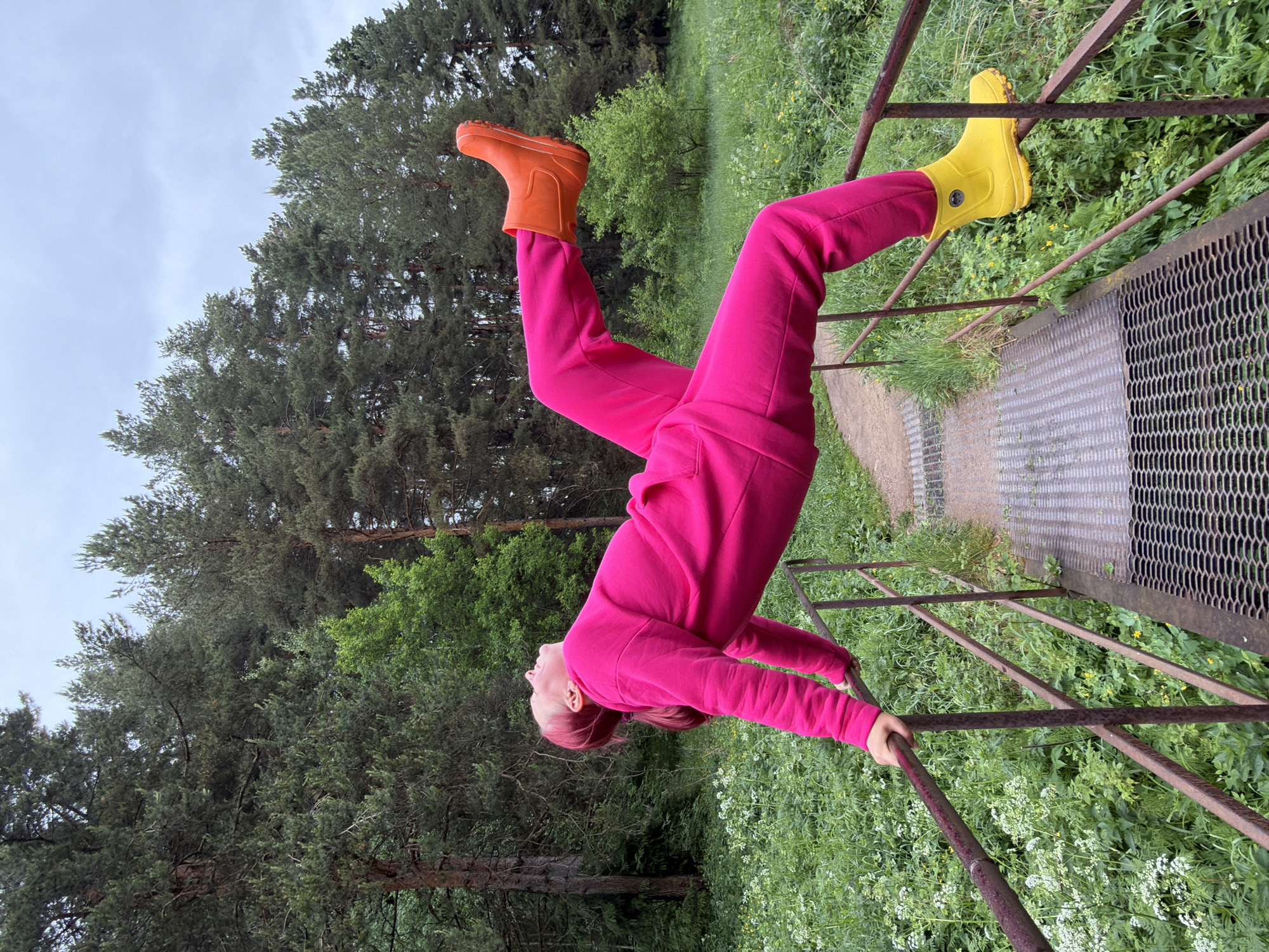 A woman in a bright pink tracksuit and mismatched orange and yellow rain boots is hanging upside down from a metal railing, presumably in a park or green space near Chaikovsky, Russia. She has red hair and is holding onto the railing with both hands, her head tilted back. The background includes trees and green foliage, as well as a metal grate walkway. The photograph is rotated.