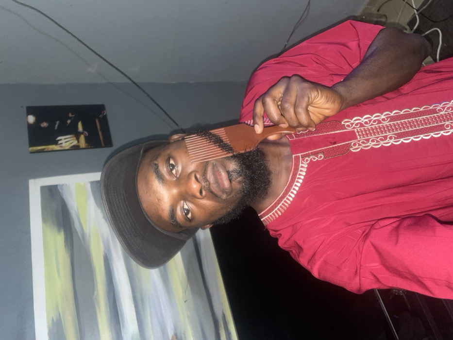 A man wearing a dark hat and red embroidered shirt holds a brown comb to his beard. He is indoors, with artwork on the wall behind him, including a painting with vertical stripes. He faces the camera, tilting his head slightly to the side. Cables are visible in the background corner. The lighting is dim.