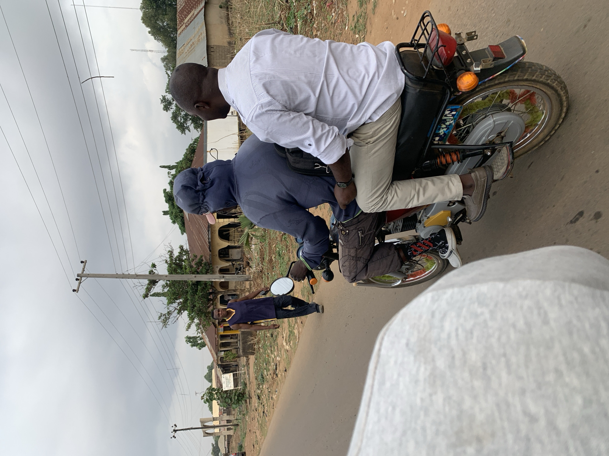 The image shows two individuals riding a motorcycle in Maigizo, Nigeria. One person wears a white striped shirt and beige pants, while the other wears a blue hooded top and dark trousers. A man in a dark blue tank top and dark jeans is walking on the side of the road. The surroundings include buildings, power lines, and a cloudy sky. A portion of the vehicle that took the picture is visible at the bottom of the frame.