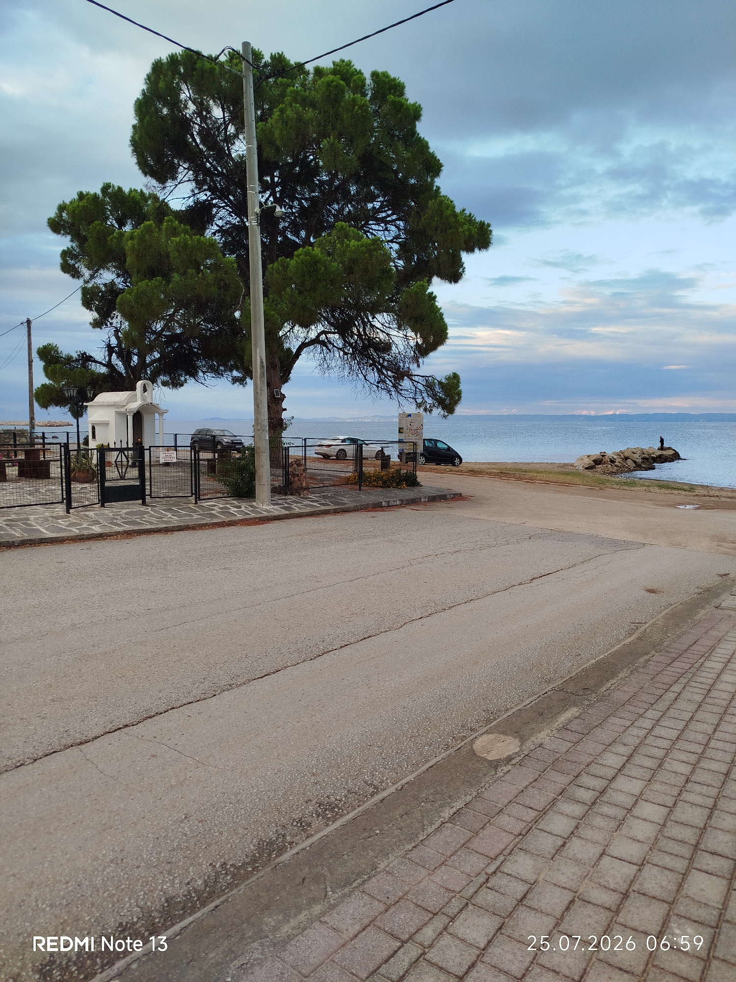 The image shows a view of the coastline in Sithonia Municipal Unit, Greece. In the foreground, there is a paved road and a sidewalk. Beyond the sidewalk, there is a small, white building resembling a chapel, surrounded by a black metal fence. A large pine tree with dark green foliage stands prominently to the left of the chapel. Several cars are parked near the chapel, and a fence separates the area from the beach. The beach itself is rocky in some areas and leads to the sea. The sea is calm, with a cloudy sky above, exhibiting hues of blue and gray. In the distance, a person is standing on a rocky outcrop by the water. The timestamp on the image reads "25.07.2026 06:59," indicating the photo was taken in the early morning. The text "REDMI Note 13" is also visible in the bottom left corner, likely indicating the phone model used to capture the image.