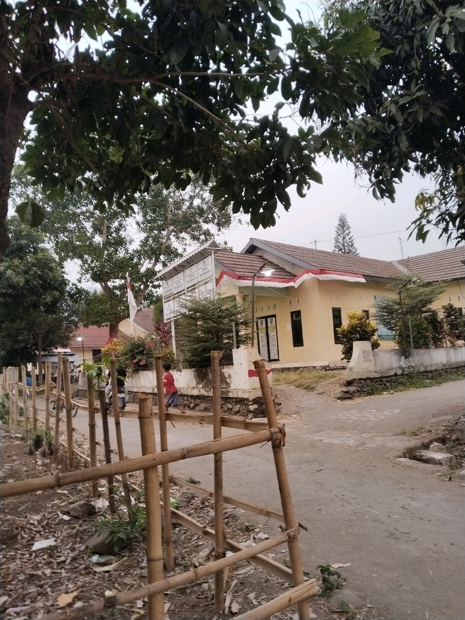 A dirt road curves around a yellow building adorned with red and white Indonesian flags. A person in a red shirt runs past the building, while another person on a motorcycle rides on the road. The building appears to be a government office in Sangkanjaya, Indonesia, with signs on the wall that are partially visible. The foreground is dominated by a bamboo fence, and the background shows trees and a cloudy sky.