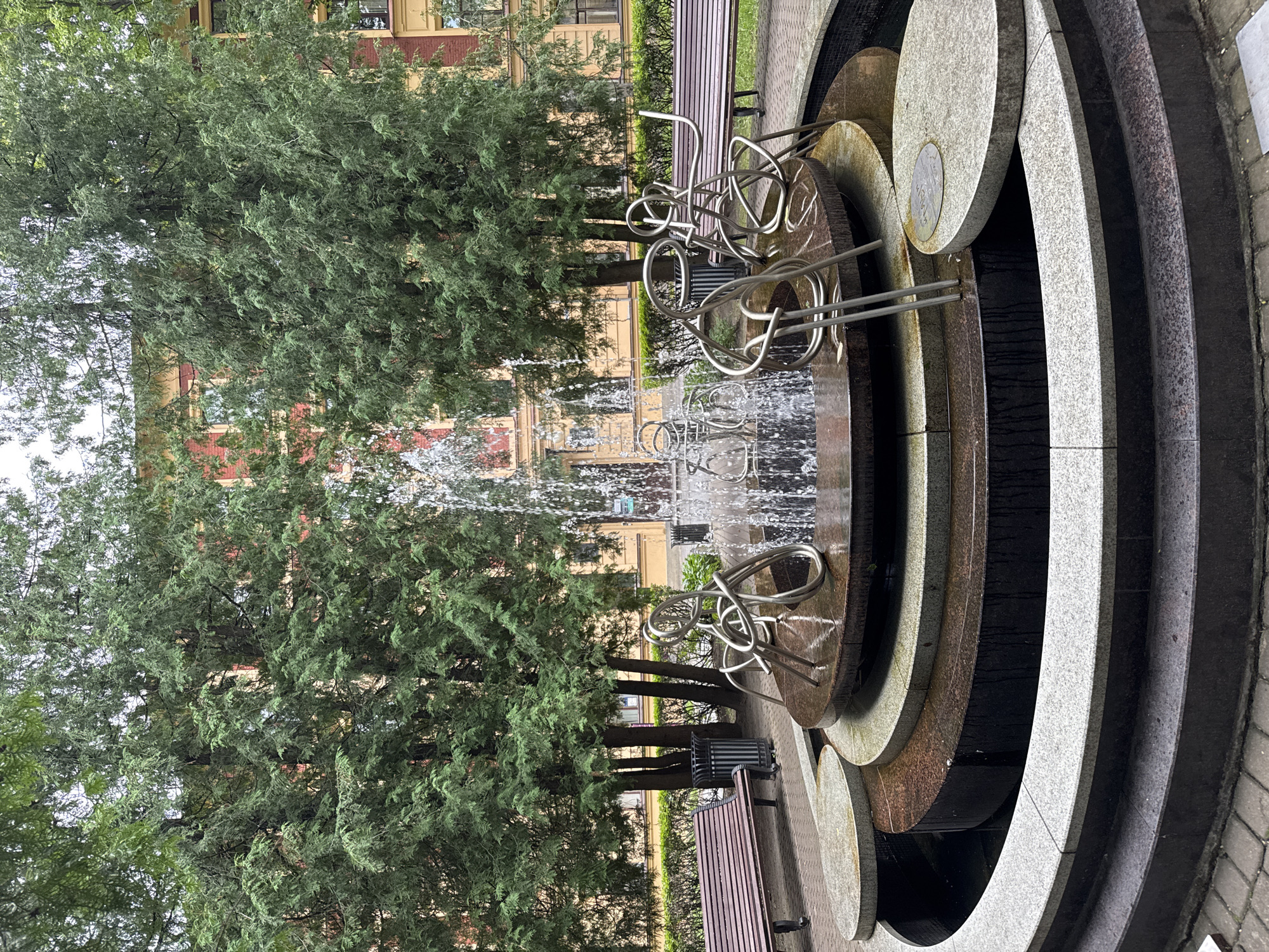 The image captures a circular fountain with a tiered stone base, located in Saint Petersburg, Russia. The fountain features abstract metallic sculptures of figures intertwined with water jets. Surrounding the fountain are lush green trees, providing a natural backdrop, and in the distance, a building with red and yellow facades is visible. Two wooden benches are positioned on either side of the fountain, suggesting a public park or courtyard setting. The ground around the fountain is paved with cobblestones. The lighting suggests it is daytime, possibly overcast.