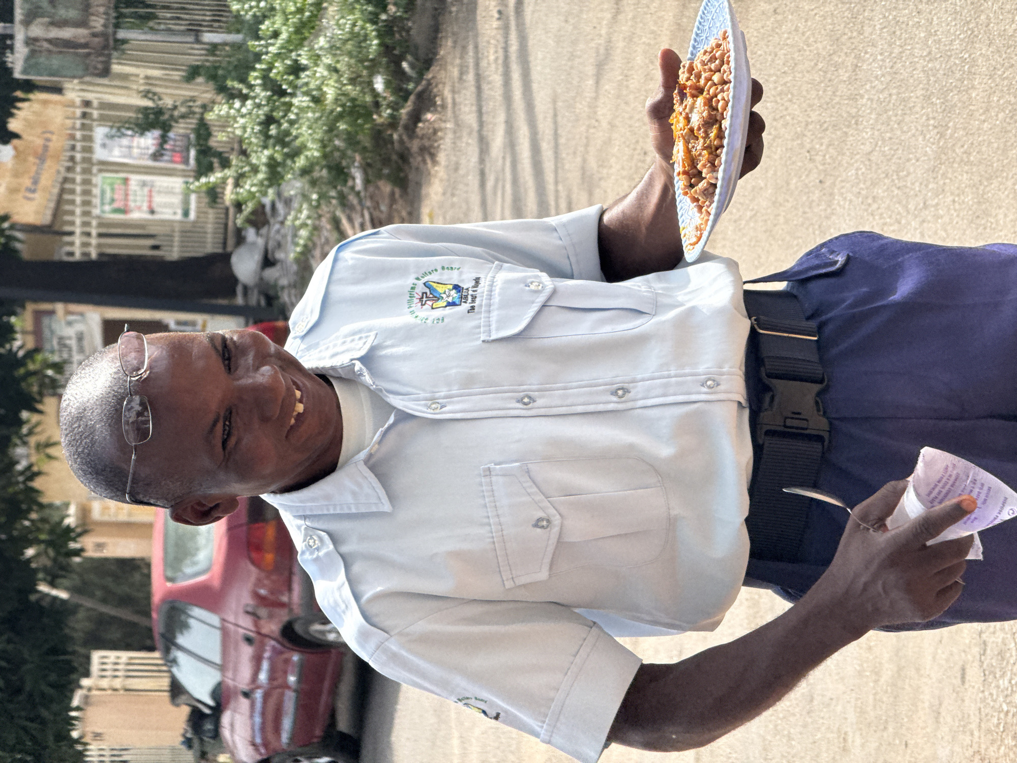 An outdoor, eye-level portrait-style shot, rotated on its side, shows a smiling Black man in what appears to be Abuja, Nigeria, holding a plate of food and a small piece of paper. He stands on a paved surface, likely a road or walkway, with buildings, a red car, and green foliage visible in the background under bright daylight.
The man, with a shaved head and glasses, wears a light blue short-sleeved collared shirt and dark blue trousers with a black belt. His shirt features a prominent circular logo on the left chest, which reads "Pilgrims Welfare Board," "ABUJA," "The Seat of Power," and "Since 1989," encompassing symbols including a crescent moon, a star, and a cross. He is looking towards the camera with a wide, friendly smile, showing his teeth.
In his right hand, he holds a light blue or white patterned plate filled with what appears to be beans and other ingredients, possibly a local dish like "beans and plantain" or a similar stew. His left hand holds a small, white, crumpled piece of paper, possibly a receipt or flyer, with some faded purple text partially visible (e.g., "PURPOSE: PROMOTE," "DISPENSE ROLLING"). A small spoon can be seen tucked into the paper.
The background includes a residential or commercial street scene. A reddish-brown car is parked behind him to his left, and further back, structures with multiple windows and balconies are visible. Green trees and shrubs flank the scene, suggesting a warm climate. The bright sunlight indicates it is daytime, and the overall mood is cheerful.