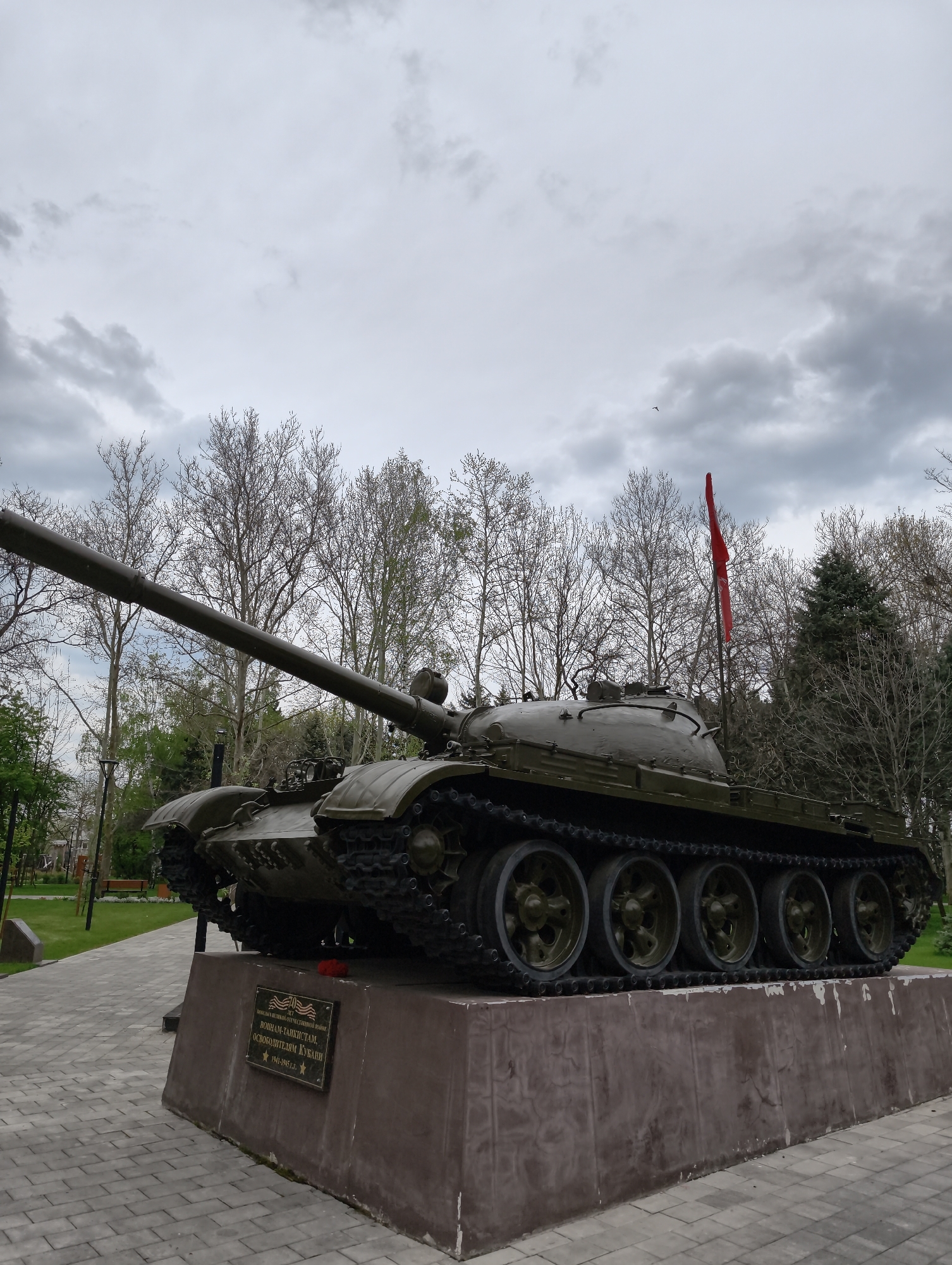 An outdoor, eye-level shot captures a military tank monument under a cloudy, overcast sky. The large, olive-green tank, likely a T-34 or similar Soviet-era model, is prominently displayed on a brown, concrete pedestal. Its long barrel extends towards the left, and its formidable tracks and road wheels are visible on both sides of the vehicle. A red flag, possibly a Soviet flag, stands mounted on the rear of the tank's turret.
The monument is situated in what appears to be a park or public square in Krasnodar, Russia. The ground surrounding the pedestal is paved with light grey interlocking bricks. In the background, numerous deciduous trees with bare branches suggest it is early spring or late autumn, while some evergreens are visible further back on the right. A small, dark bird is captured mid-flight in the upper right portion of the sky.
On the front of the pedestal, a dark plaque with gold or brass lettering is affixed. A small red flower or wreath rests on the pedestal directly beneath the plaque. The text on the plaque, in Cyrillic, reads:
"ВЕЛИКОЙ ОТЕЧЕСТВЕННОЙ ВОЙНЫ" (of the Great Patriotic War)
"ВОИНАМ-ТАНКИСТАМ, ОСВОБОДИТЕЛЯМ КУБАНИ" (To the tank-soldiers, liberators of Kuban)
"1941-1945 г.г." (1941-1945 years)
Below the dates is a five-pointed star emblem. The overall scene conveys a solemn and commemorative atmosphere.