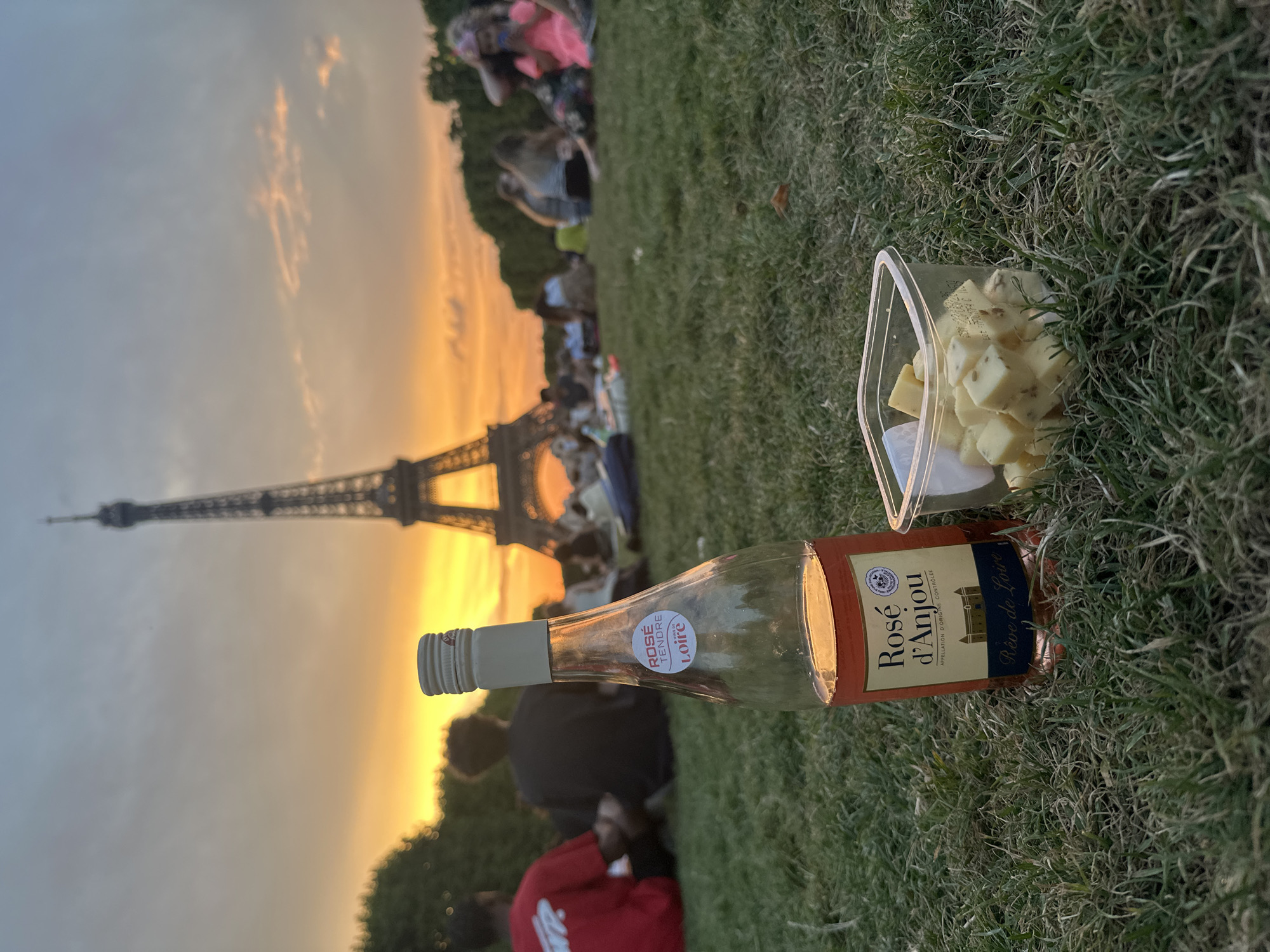 In Paris, France, on a grassy field in front of the Eiffel Tower during sunset, a bottle of Rosé d'Anjou wine and a clear plastic container filled with cubed cheese are placed on the grass. The Eiffel Tower is visible in the background, towering over a crowd of people sitting on the lawn. The sky is painted with warm hues of orange and yellow from the setting sun, with scattered clouds. Several individuals are casually gathered on the grass, some sitting and others standing, in the mid-ground and background. The foreground focuses on the bottle of wine and the container of cheese, suggesting a picnic or leisurely outdoor gathering.