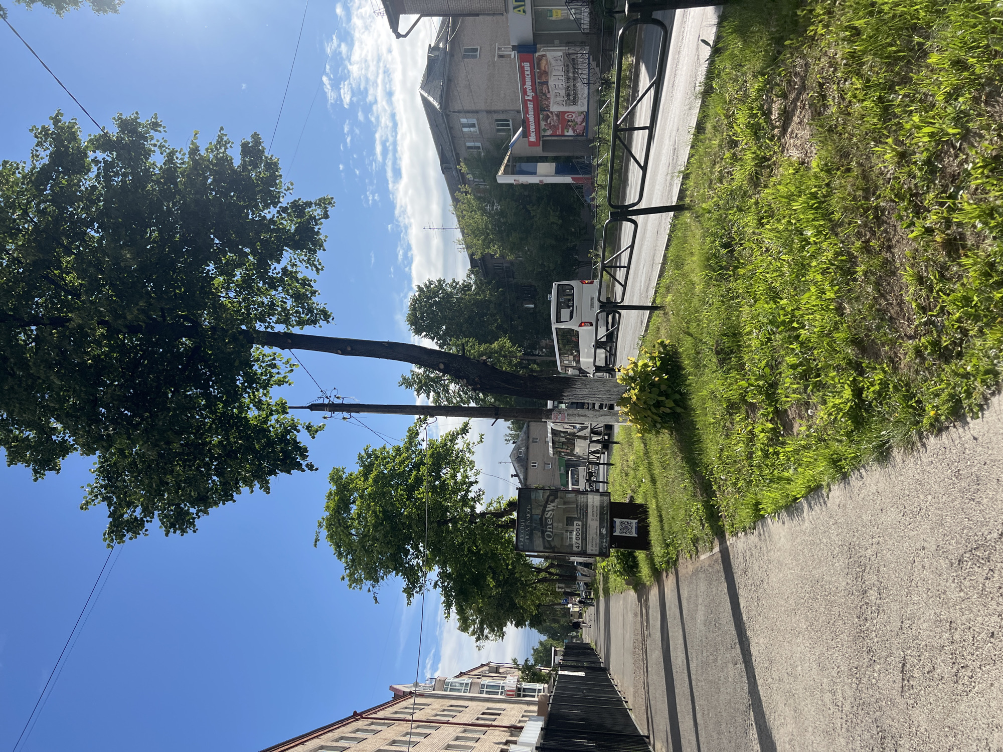 An eye-level, medium shot captures a sunlit street scene in Chaikovsky, Russia. A paved sidewalk runs along the left side of the image, bordered by a black metal fence and a multi-story brick building with several windows. Beyond the fence, a road is visible with a black metal railing separating it from a grassy verge. Several trees line the street, their lush green leaves casting shadows. A few people are walking on the sidewalk in the distance. On the right side of the road, a white bus is seen, and further down, a building with signage that appears to be a shop. The sky is bright blue with scattered white clouds. The overall atmosphere is that of a clear, sunny day in a residential urban area.