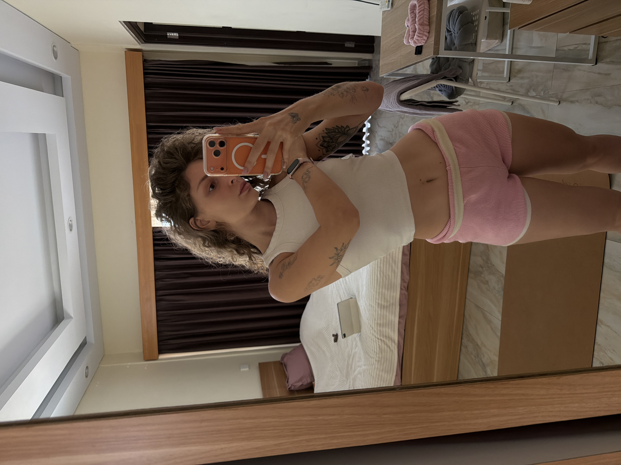 A woman is taking a mirror selfie in what appears to be a bedroom in North Kuta, Indonesia. She is wearing a white crop top and pink ribbed shorts. Her hair is curly and she has tattoos on her arms. In the background, there is a bed with a purple pillow and a tablet on it. To the right of the bed, there is a desk with chairs and various items on it, including a pink headband. The room has brown curtains and a wooden framed mirror. The floor is tiled and there is a brown mat visible.