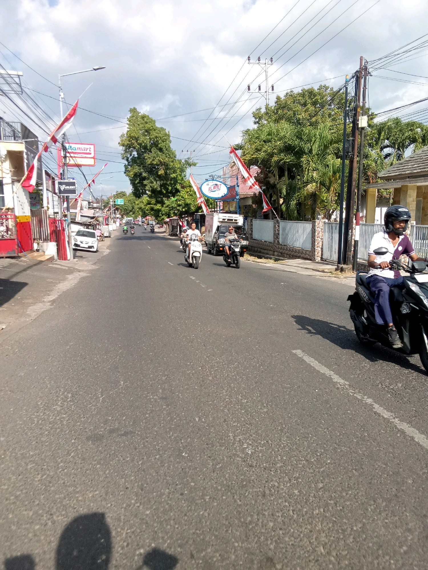 This is a full-frame, eye-level, outdoor, daylight, medium-shot photograph taken from a moving vehicle on a paved road in Gebang, Indonesia. The road is a two-lane asphalt road with a white dashed line dividing the lanes. The road is lined with buildings and trees on both sides, and utility poles and wires are visible overhead. Several people are riding motorcycles, with some in motion and some stopped. One person, a man wearing a helmet, a white and purple polo shirt, and dark pants, is prominently featured riding a black motorcycle in the foreground, moving from left to right. Other individuals on motorcycles are visible in the background, some of whom are wearing helmets. Flags, appearing to be the Indonesian flag, are displayed on poles along the street. Signs for businesses such as "Indomaret" and "Planet" are also visible. The sky is partly cloudy with blue sky visible. The overall scene depicts a typical street view in an Indonesian town, with local traffic and commercial establishments.