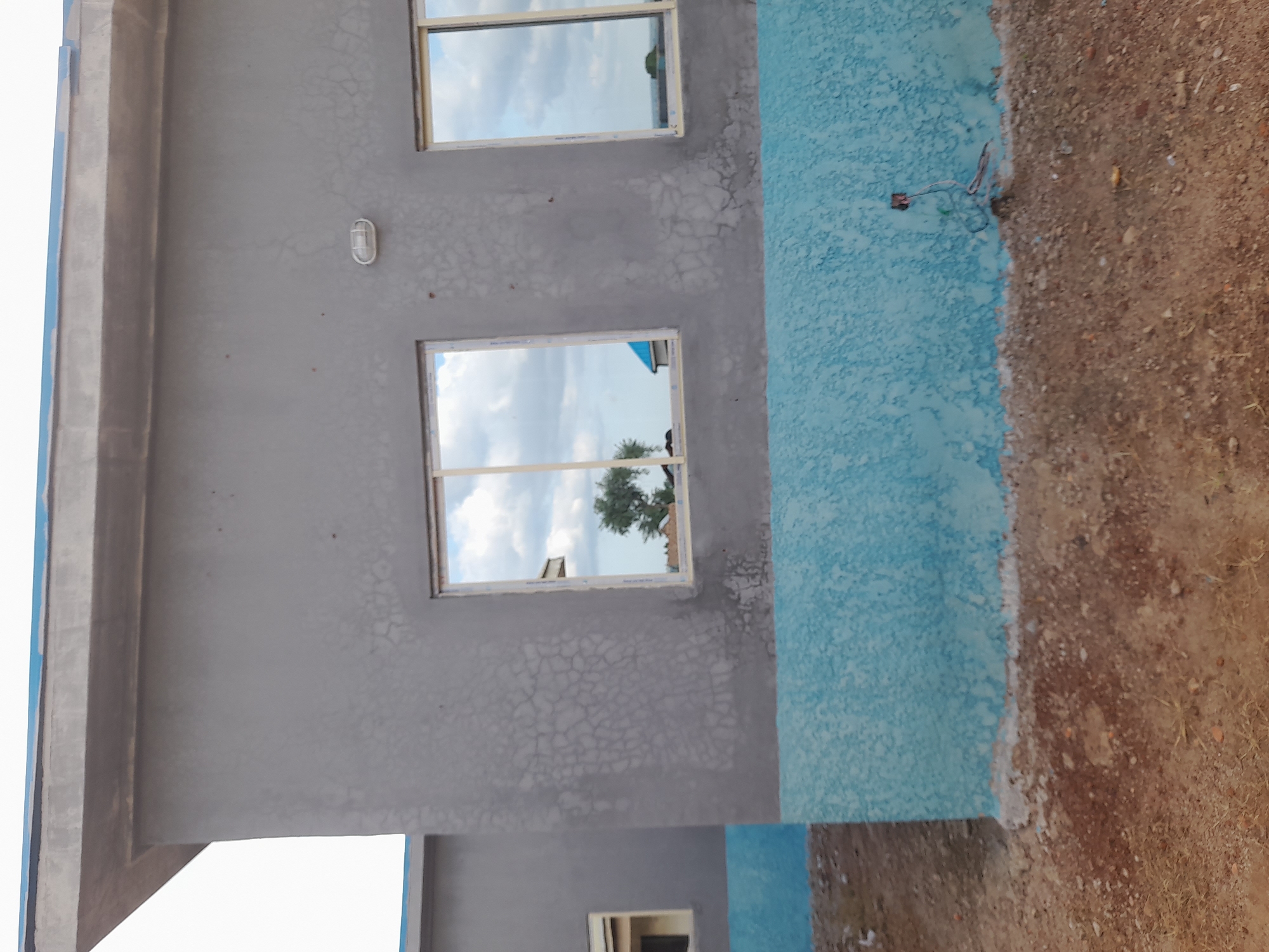 The image shows a side view of a building under construction in Ibi, Nigeria. The building has gray walls with a lighter gray textured pattern. There is a light blue band across the lower portion of the walls. Two windows are visible on the right side of the building, with reflections of clouds and trees. A small, rectangular light fixture is mounted on the wall between the windows. To the left, another building or section of the same building is visible, also with a light blue lower band and a small window. The ground in front of the building is rough and consists of dirt, gravel, and some dry grass. There is a small electrical box with wires protruding from it near the base of the right section of the building. The sky is visible at the top and is cloudy.