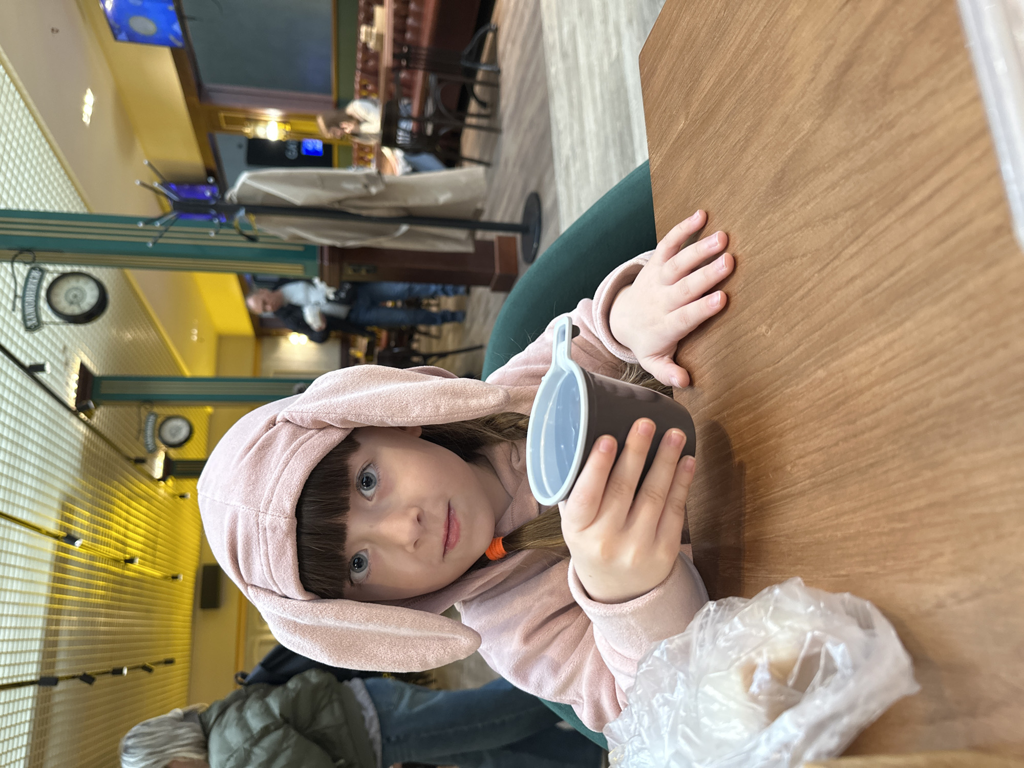 A young child wearing a pink bunny-ear hoodie is seated at a wooden table in an indoor public space, possibly a restaurant, in Bolshoye Savino, Russia. The child is holding a brown and white cup and has a plastic bag nearby. The environment includes interior architectural details like a patterned wall and visible features of a public establishment, such as tables, chairs, and overhead lighting. Other individuals are present in the background, some seated and others standing or walking. A clock is also visible. The scene appears to be a casual, everyday moment.