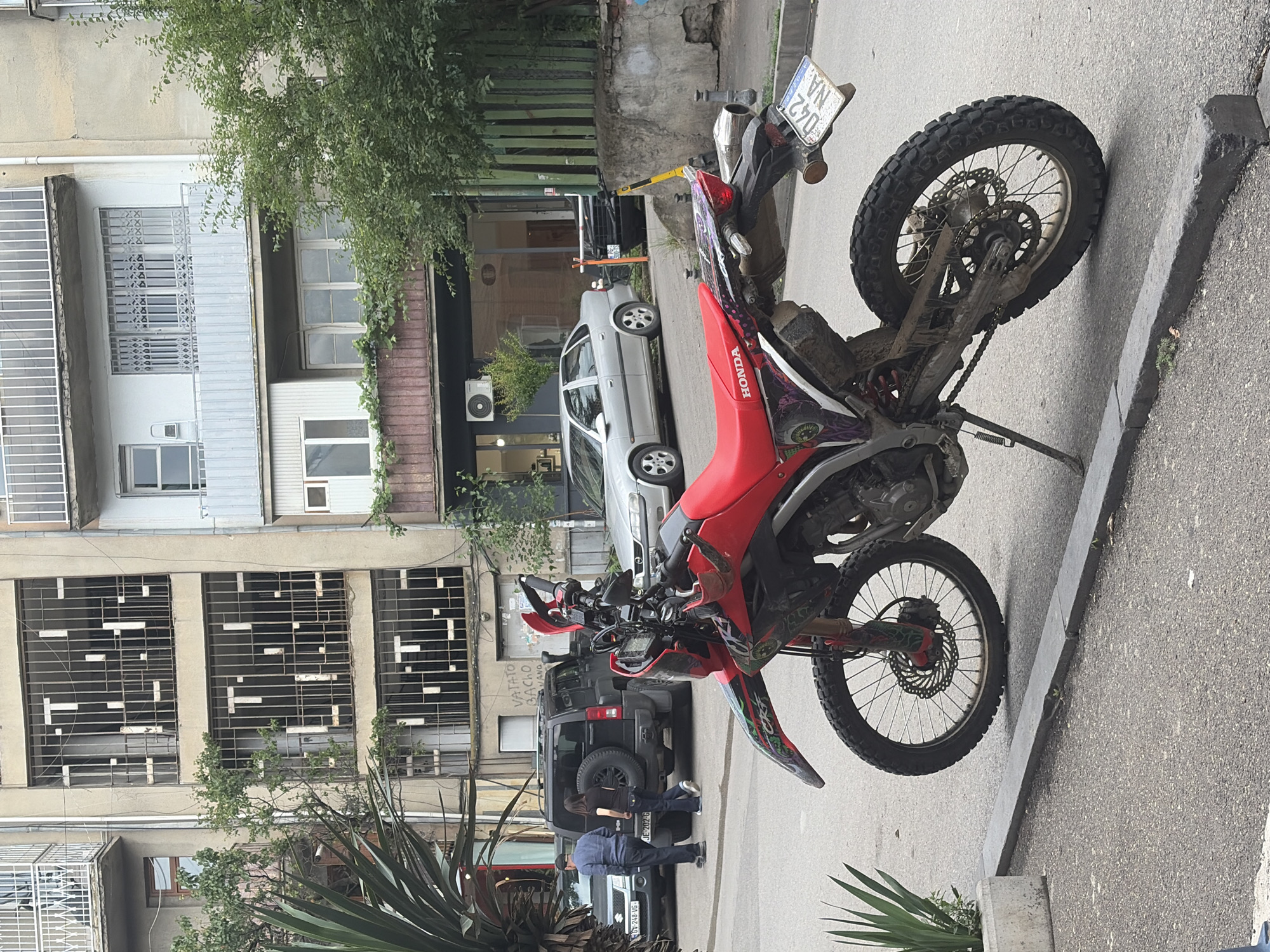A red Honda dirt bike is parked on a street in Tbilisi, Georgia, with its kickstand down. The bike is positioned at an angle, facing towards the right of the frame, with its front wheel slightly elevated. The bike's seat is red, and it has a license plate that reads "042 NA." The background shows a multi-story building with balconies and windows, as well as trees and other parked vehicles, including a black SUV and a silver sedan. The street appears to be paved, with a concrete curb in the foreground. The overall scene suggests an urban environment.