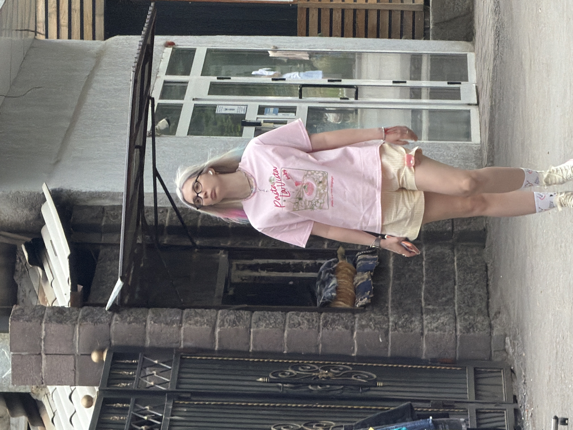 A young woman with blonde hair with pink streaks and glasses is walking on a paved surface in front of a building. She is wearing a pink t-shirt with a graphic print and the words "Pasta La Vista, baby", light yellow shorts with a red detail on the side, and white socks with a pattern. She has a red bracelet on her left wrist and is holding a phone in her right hand. In the background, there is a dark metal fence with ornate details on the left, and a building with large glass doors and windows on the right. A portion of the building is covered with horizontal wooden planks. The ground is a mixture of concrete and dirt.