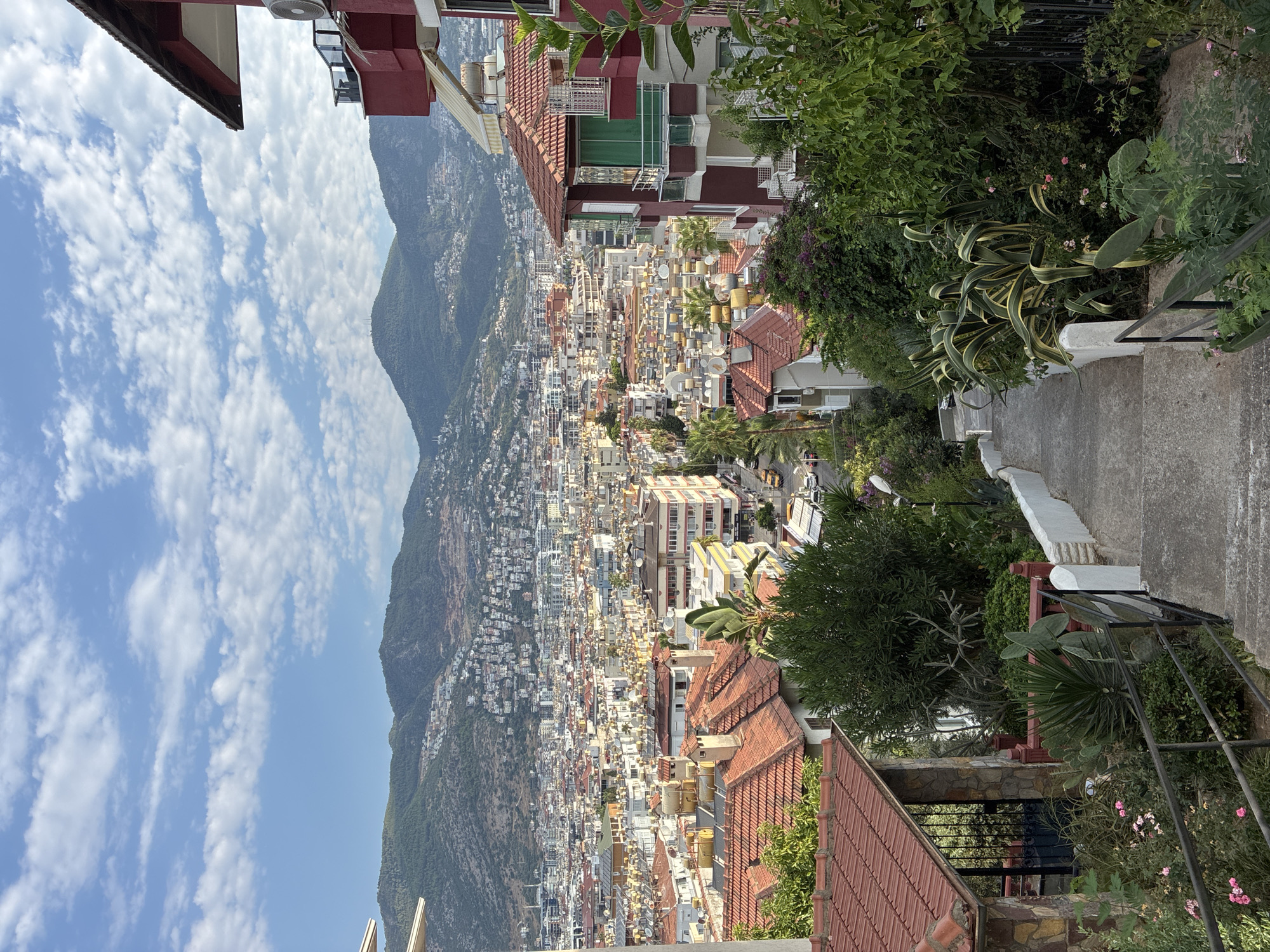 A high-angle shot captures a cityscape in Alanya, Turkey, with a focus on the layered buildings and mountainous backdrop. In the foreground, a concrete staircase with metal railings descends, flanked by lush greenery and flowering plants. A small stone structure with a metal gate is visible on the left.
The midground reveals a dense urban environment characterized by numerous buildings with red-tiled roofs, many equipped with solar water heaters. A street lined with cars and palm trees runs through this area.
The background is dominated by a sprawling city that climbs the slopes of a verdant mountain range. The sky above is a vibrant blue, dotted with scattered white clouds. The overall scene depicts a sunny day in a coastal city, characterized by its hilly terrain and architectural density. No people are visible in the image.