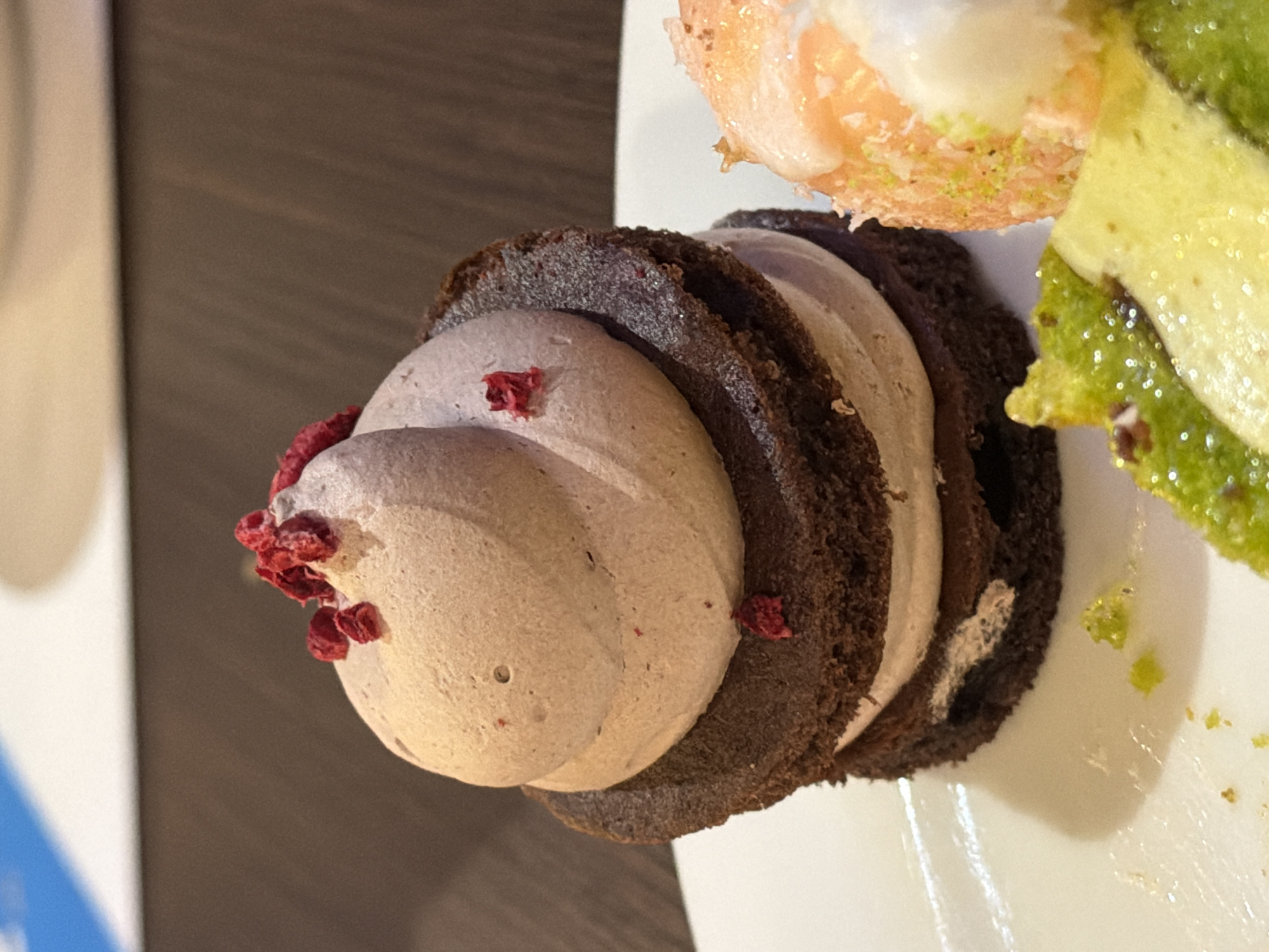 The image shows a close-up of desserts on a white plate. The main subject is a layered dessert, featuring dark, possibly chocolate cake-like rounds interspersed with a light-colored filling, topped with a pinkish-gray cream and what appears to be dried raspberry pieces. Adjacent to it, partially visible, is another dessert featuring a light orange and white layer next to a green layer. The plate rests on a dark wooden surface, and a glimpse of a blue object is seen on the left. The dessert appears to be indoors and likely on a table. While it's impossible to pinpoint the exact location within Varna, Bulgaria, based solely on this image, the desserts suggest it could be in a cafe, restaurant, or bakery in the city.