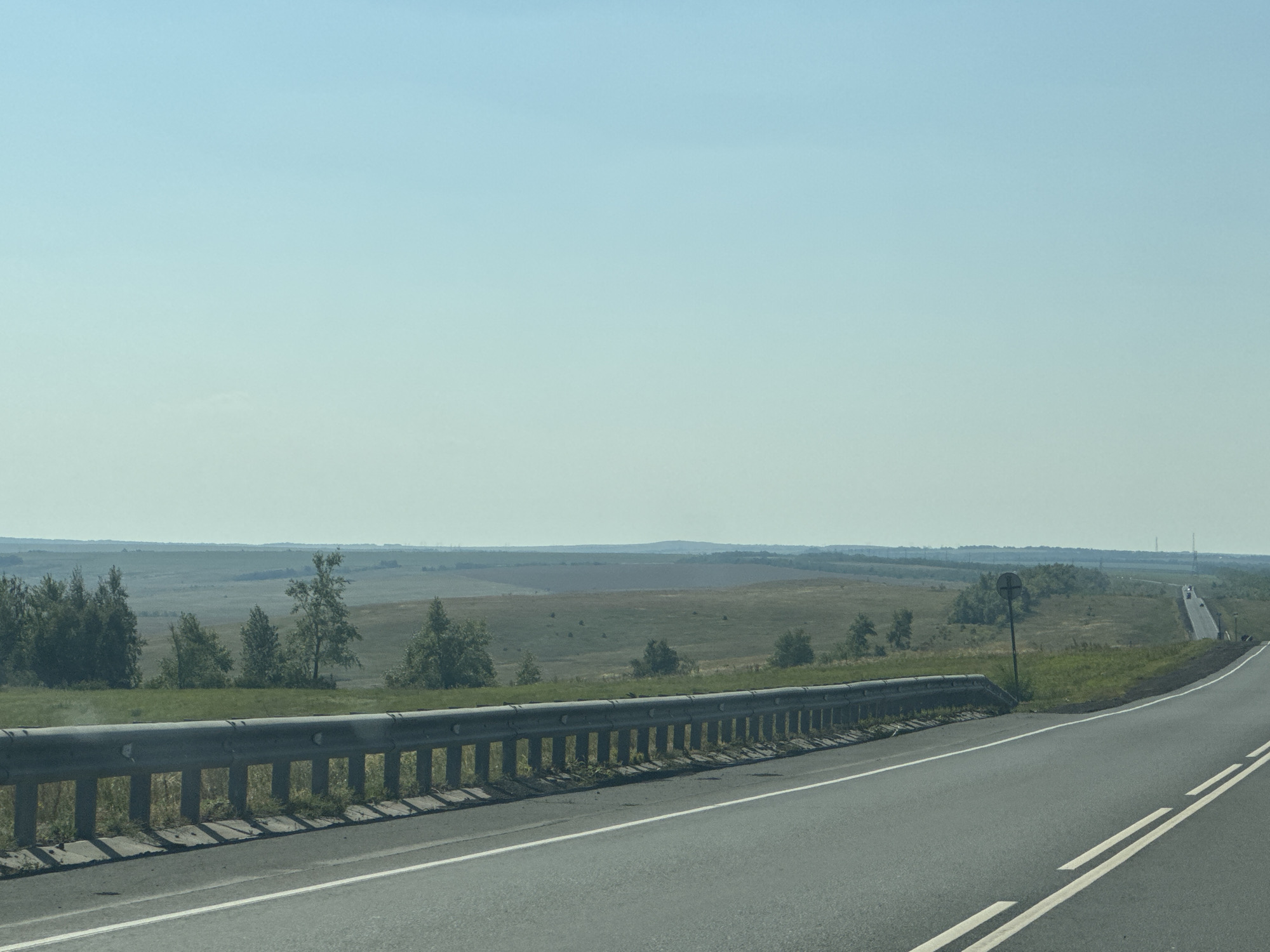 The image shows a highway in a rural area with a guardrail on the left side of the road. The road has two lanes and white markings. In the background, there are fields, trees, and rolling hills under a clear blue sky. There is a road sign visible in the distance. The scene is captured from a moving vehicle. No people are visible in the image.