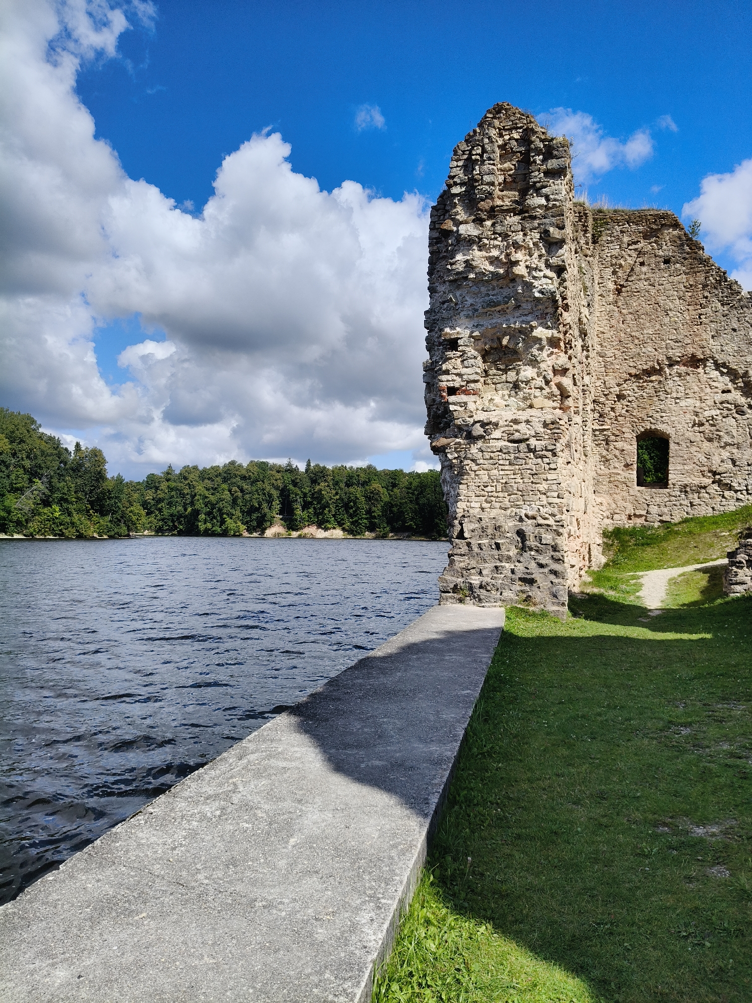 The image shows the ruins of the Koknese Castle in Koknese, Latvia. The ruins are made of stone and are located next to a body of water, likely the Daugava River. The sky is blue with some white clouds. There is green grass and trees in the background. The image appears to be taken on a sunny day.
