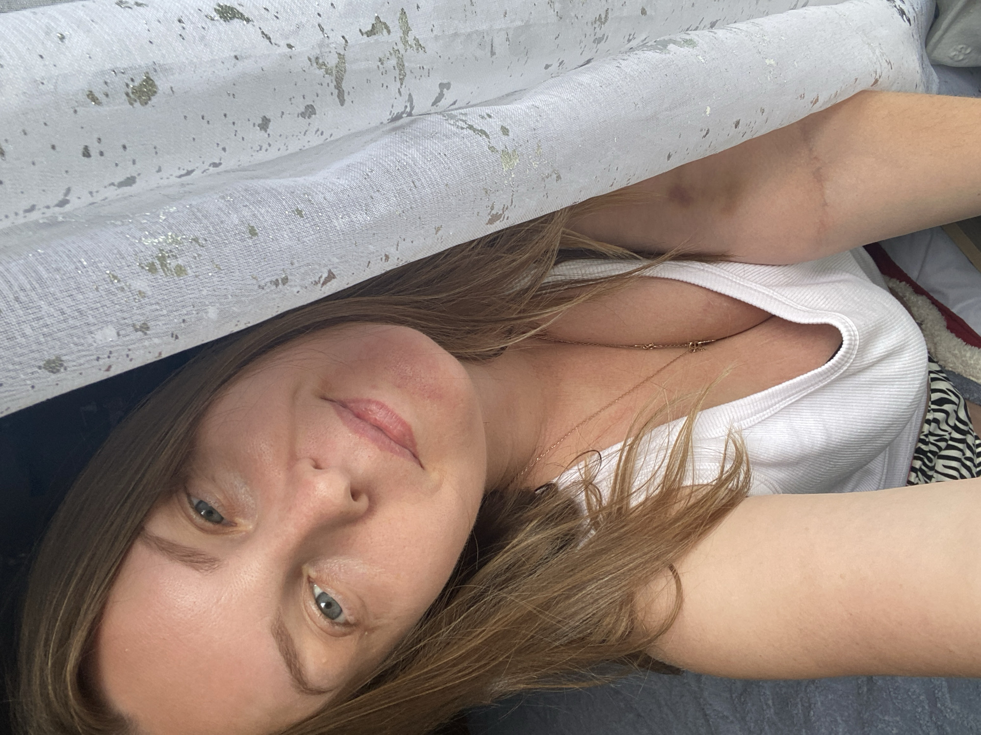 The image shows a close-up, angled selfie of a woman indoors. She has light skin, light brown hair, and blue eyes. She is wearing a white ribbed tank top and a gold necklace with a cross pendant. A white curtain with gold flecks is to her right. The background appears to be a bed with a grey and white striped blanket. The woman is looking towards the camera with a slight smile. The image appears to have been taken in an indoor setting, possibly a bedroom or living room. There is no identifiable information in the image to determine the specific location in Noviy, Russia.
