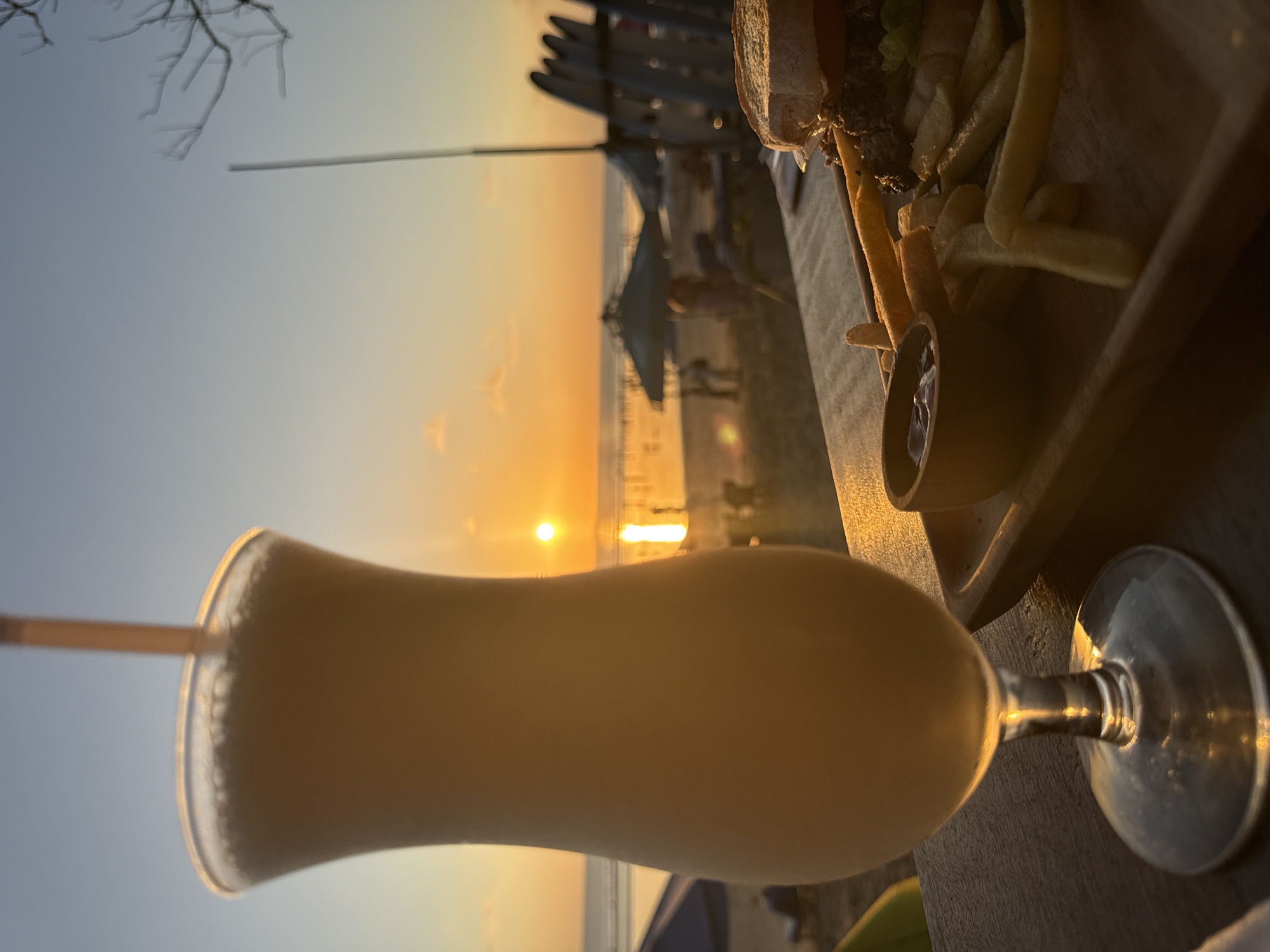 A close-up, waist-level shot captures a tall, opaque, light-colored beverage in a stemmed glass on a dark wooden surface. A straw extends from the top of the drink, and the glass is illuminated by a warm, golden light. In the background, a beach scene unfolds at sunset. The sun is a bright, orange orb low on the horizon, casting a shimmering reflection on the ocean. Silhouettes of people are visible on the beach and in the water, along with beach umbrellas and chairs. To the right of the drink, a wooden tray holds a burger, french fries, and a small bowl of ketchup. Surfboards are stacked vertically in the far right background. The overall impression is a relaxed evening meal by the sea, likely in Kuta, Indonesia, known for its sunset views and beachside dining.