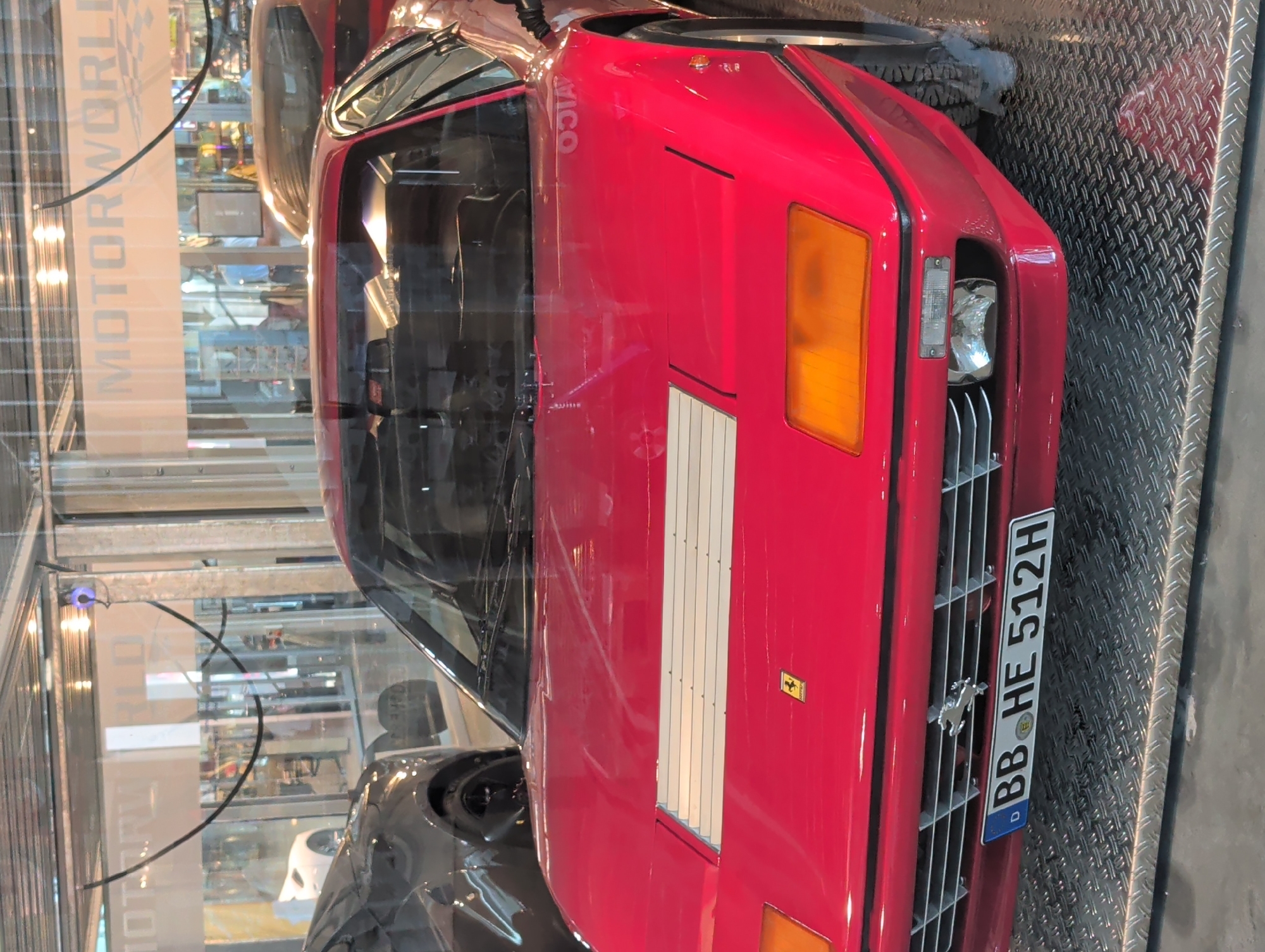 This is a close-up, slightly angled shot of the front of a bright red, classic sports car, likely a Ferrari, parked indoors. The car is positioned diagonally in the frame, with its front grille, headlights, and part of the hood prominently displayed. The car has a distinctive rectangular orange turn signal on the passenger side and a clear headlight with a chrome reflector. The grille features horizontal bars, and a chrome emblem, possibly a prancing horse, is visible.
Attached to the front grille is a license plate with white text on a black background, reading "BB HE 512H" and a blue European Union strip on the left. The car is parked on a textured metal surface, possibly a diamond plate. In the background, through a glass partition, a blurry scene of a showroom or exhibition hall is visible, with displays and lighting suggesting a public venue. A sign with vertical text "MOTORWORLD" is partially visible on the left. The lighting suggests it is daytime, and the indoor setting indicates controlled environmental conditions. There are no people visible in the frame. The overall impression is of a well-maintained classic vehicle on display. The location context of Böblingen, Germany, aligns with the "BB" on the license plate, which often indicates the district.