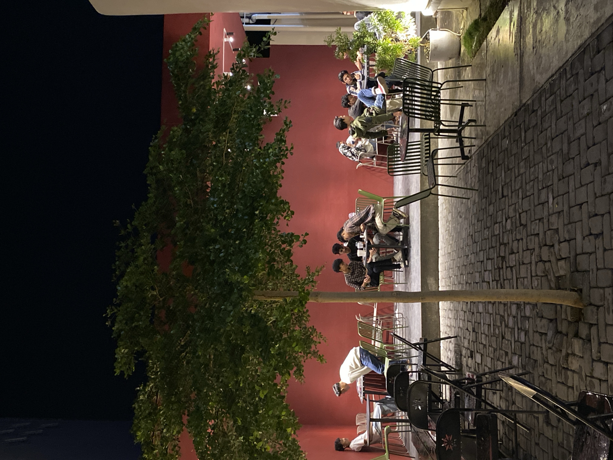 The image depicts a vibrant nighttime scene at an outdoor cafe or patio in Lhokseumawe, Indonesia. The setting features a prominent, smooth red wall as a backdrop on the left side, contrasting with a dark sky that indicates late evening or night. On the right, the ground is paved with dark, rectangular cobblestone-like bricks, suggesting a pathway or extended seating area.
Several groups of young men are seated at tables, engaged in conversation and socializing. They are casually dressed, occupying various metal chairs and tables. Some chairs are standard black slatted designs, while others have lighter frames, including some with green accents. In the foreground and extending towards the middle, a lush green tree with dense foliage partially obscures the red wall, adding a natural element to the urban environment. Another thinner tree trunk rises from the paved ground, acting as a subtle divider.
Artificial lighting, likely from overhead fixtures and possibly spotlights, illuminates the scene, casting bright glow on the people and parts of the red wall, creating pockets of light and shadow. A few potted plants with greenery are also visible near the paved area. The overall atmosphere appears relaxed and social, with individuals enjoying each other's company in the cool evening air. No discernible text is visible within the frame.