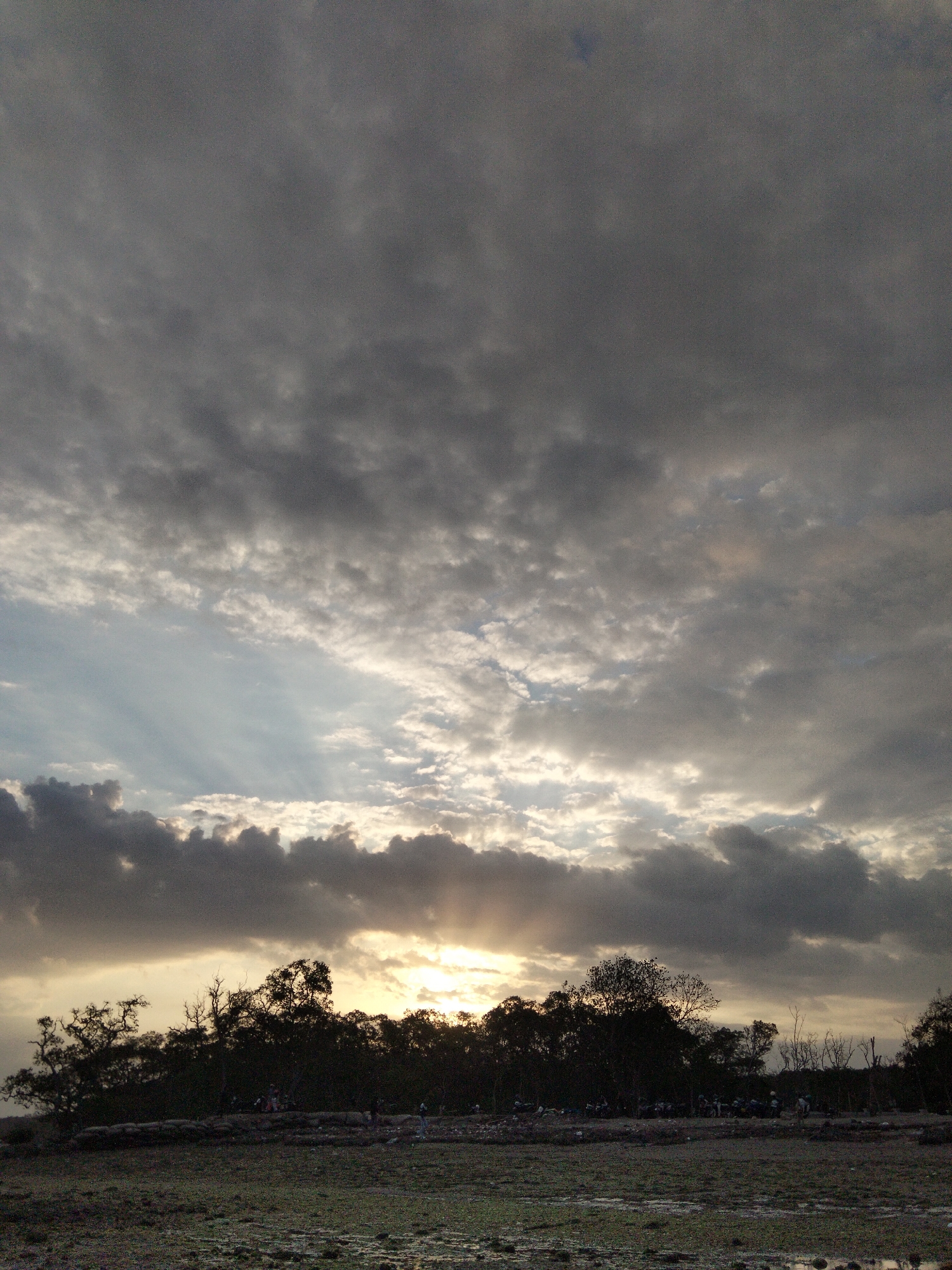 The image depicts a sunset scene over a dense treeline in an unknown location in Indonesia. The sky is filled with dramatic, heavy clouds, with rays of sunlight breaking through in the center. Below the treeline, which is silhouetted against the bright sky, a muddy, marshy area is visible, with scattered debris and what appear to be a few people and motorcycles in the distance. The foreground is a textured, muddy ground. The overall mood is somber and atmospheric, capturing the raw beauty of nature at dusk.