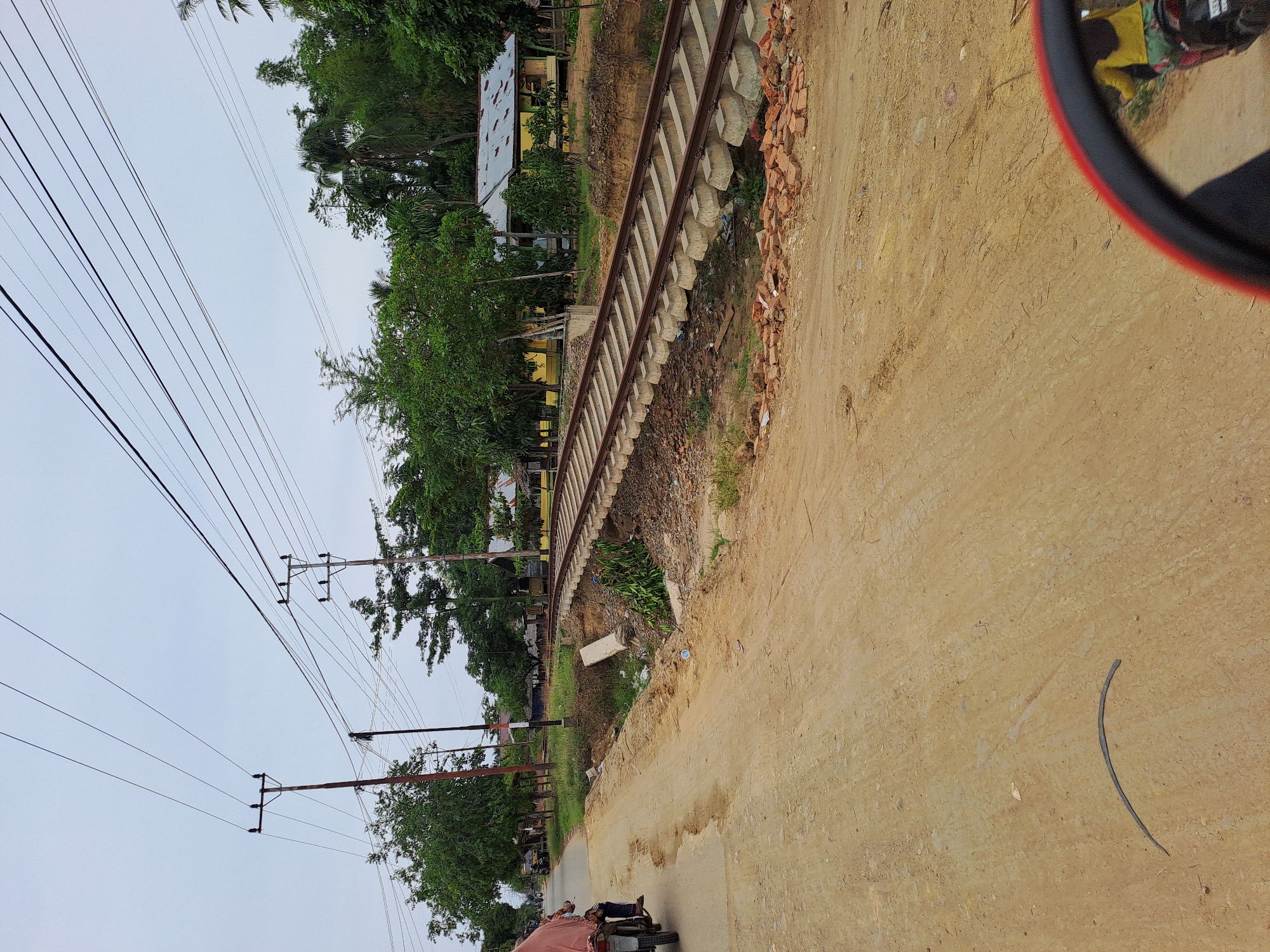 In the city of Geulumpangsulu Timu, Indonesia, a dirt road curves to the left, meeting a paved road at an intersection. Overhead, power lines stretch across a cloudy sky, supported by wooden poles. To the right of the paved road, a railway track is elevated on concrete sleepers, spanning a shallow ditch filled with debris and vegetation. Beyond the tracks, houses with corrugated metal roofs and yellow walls are visible amongst lush green trees. On the dirt road, a person rides a motorcycle with a passenger, both covered in a draped cloth. The scene is illuminated by diffused daylight, suggesting an overcast day.
