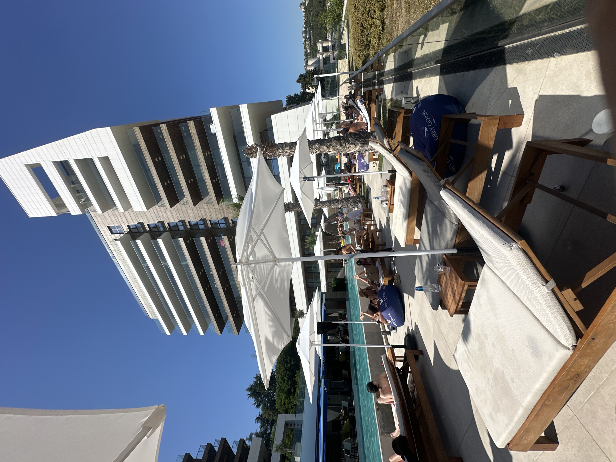 This image captures a sunny day at an outdoor swimming pool area, likely a resort or a high-end establishment, with a modern architectural building in the background. The foreground features several empty lounge chairs on a tiled patio, some with plush cushions, indicating a place for relaxation and sunbathing. To the left, a large white umbrella provides shade over a portion of the patio. Beyond the chairs, a bright blue swimming pool is visible, with several people relaxing by its edge. Some individuals are seated on beanbag chairs, while others lounge on beds by the pool. A bar or restaurant area with blue awnings is situated behind the pool. The architecture of the building in the background is striking, with multiple levels of balconies and a unique, angled white facade. The sky is a clear, vibrant blue. The overall scene suggests a luxurious and leisurely atmosphere.