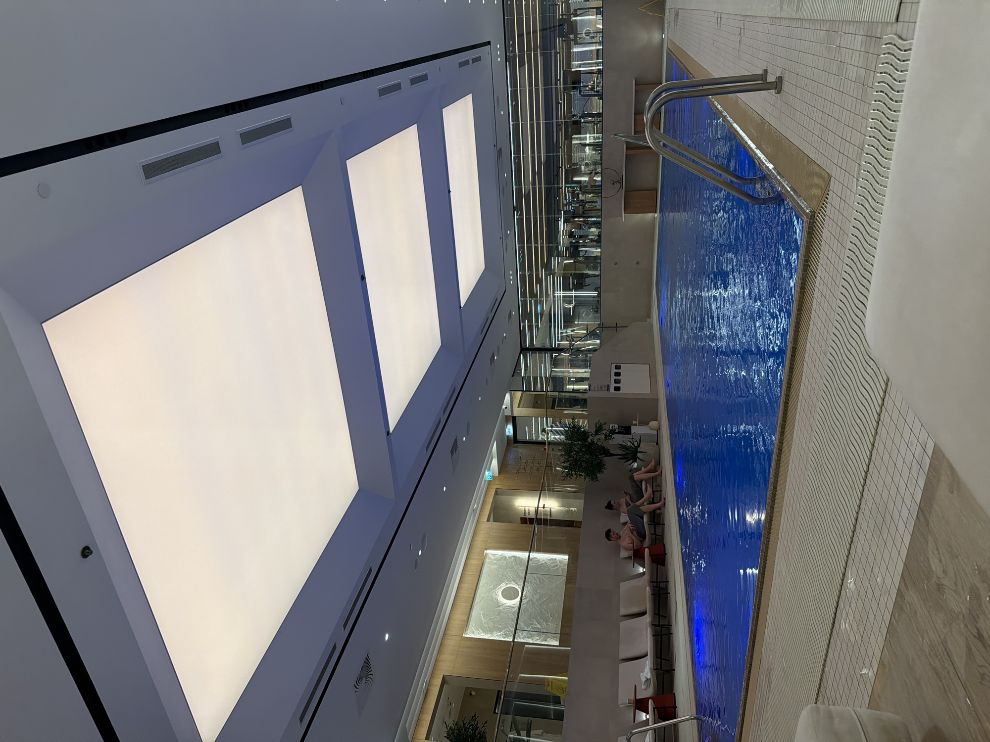 The image shows an indoor swimming pool area. The pool itself is rectangular with bright blue water, and a stainless steel ladder is visible at one end. Next to the pool, there are several lounge chairs, some of which are occupied by people relaxing.
Above the pool and lounge area, there's a high ceiling with a series of large, rectangular light panels illuminating the space. The overall impression is a modern and well-lit recreational facility. The location is unknown.