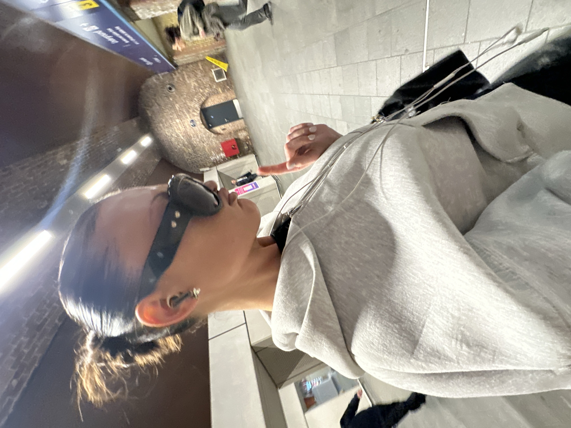 The image depicts a scene inside London Bridge Underground station in London, United Kingdom. In the foreground, a person is visible from the chest up, in profile facing right. This individual has dark hair pulled back into a bun, wears large dark sunglasses, a small light-colored earbud in the left ear, and a stud earring below it. They are dressed in a light grey hooded sweatshirt with a zipper, over a dark garment, and a thin wire necklace is visible. Their right hand is raised, with the index finger pointing upwards, revealing light-colored nail polish. A black bag is partially visible on their left side.
In the mid-ground, to the far left, another person in dark clothing is partially visible. Further into the background, a third person is walking or running away to the right, wearing a light-colored jacket, dark trousers, and carrying a dark backpack.
The environment features distinctive reddish-brown brick archways and an arched ceiling with multiple linear fluorescent light fixtures. The floor is covered with light grey square tiles. On the right side of the background, a blue overhead sign with white text and an arrow points right, indicating "Underground" and other directions. A dark door is set within a central brick archway. To its left, a red fire extinguisher box is mounted on the brick wall, and to its right, a yellow caution cone and a white rectangular object are visible on the floor and wall respectively.