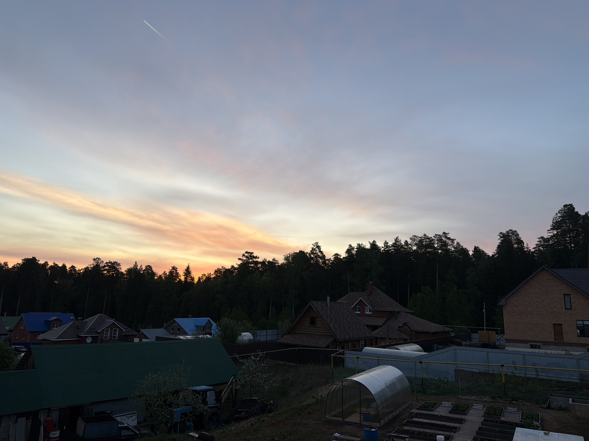 The image depicts a residential area near a forest in the city of Chaikovsky, Russia, during a colorful sunset. The sky shows bands of pink, orange, and blue hues. In the foreground, various houses with differently colored roofs (green, blue, brown) and construction materials are visible. There are multiple greenhouses on the right side of the frame, along with garden beds. A dense forest forms the backdrop, separating the residential area from the horizon. An airplane contrail can be seen in the upper left portion of the sky. The scene appears still and quiet.